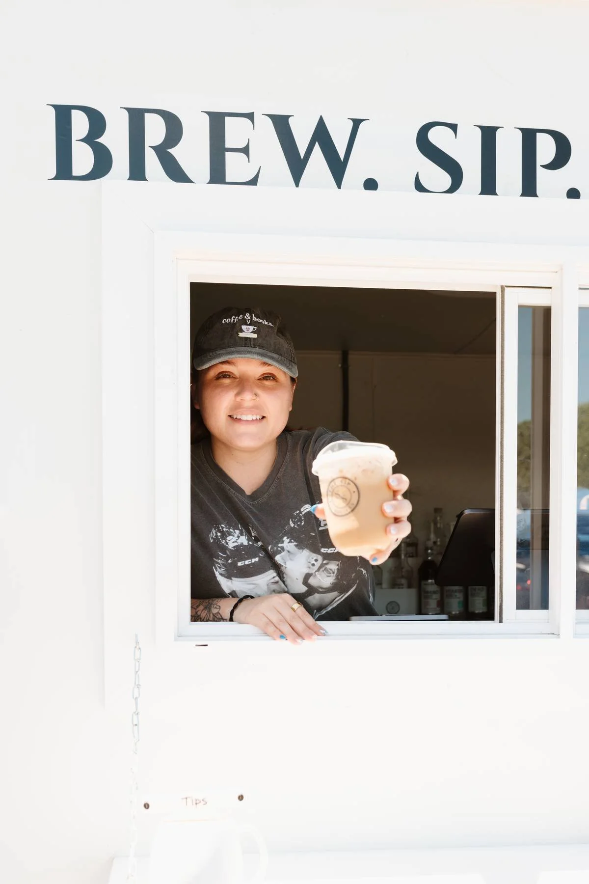 A woman smiling and holding a cup of iced coffee outside a coffee stand with the sign 'BREW.SIP.'