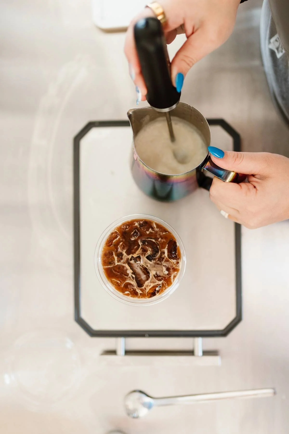A person with blue nail polish and a gold ring on their finger is blending a drink in a stainless steel pitcher. In front of them is a cup of iced coffee with ice cubes and milk, on a tray.
