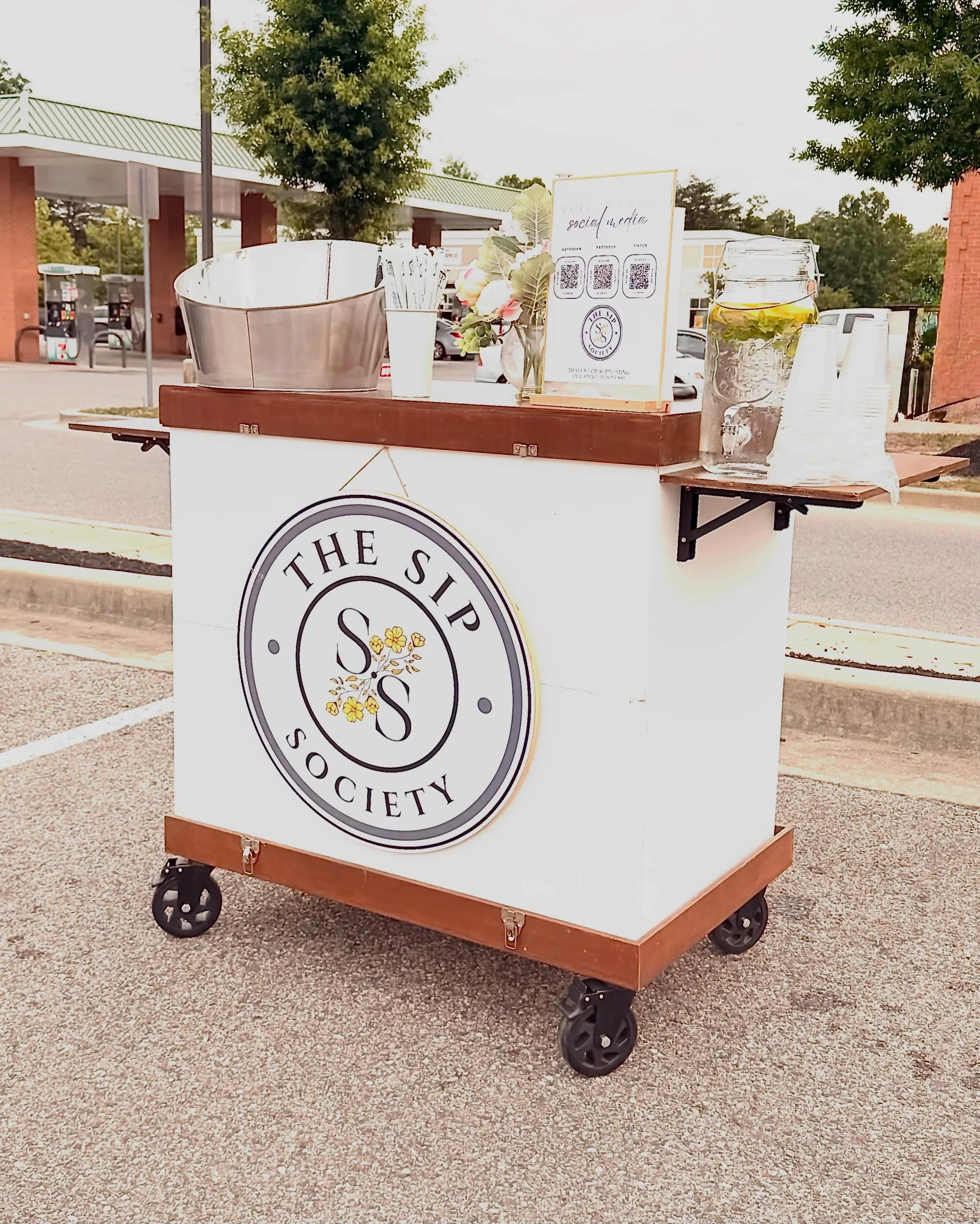 A mobile cart with a white front panel displaying a logo for The Shipp Society with black, gray, and yellow design elements; the cart has four black wheels and a wooden shelf on top holding various items, including a large metal bowl, cups, a jar with lemon and mint slices, a sign with QR codes and social media information, and decorative flowers in a white vase.
