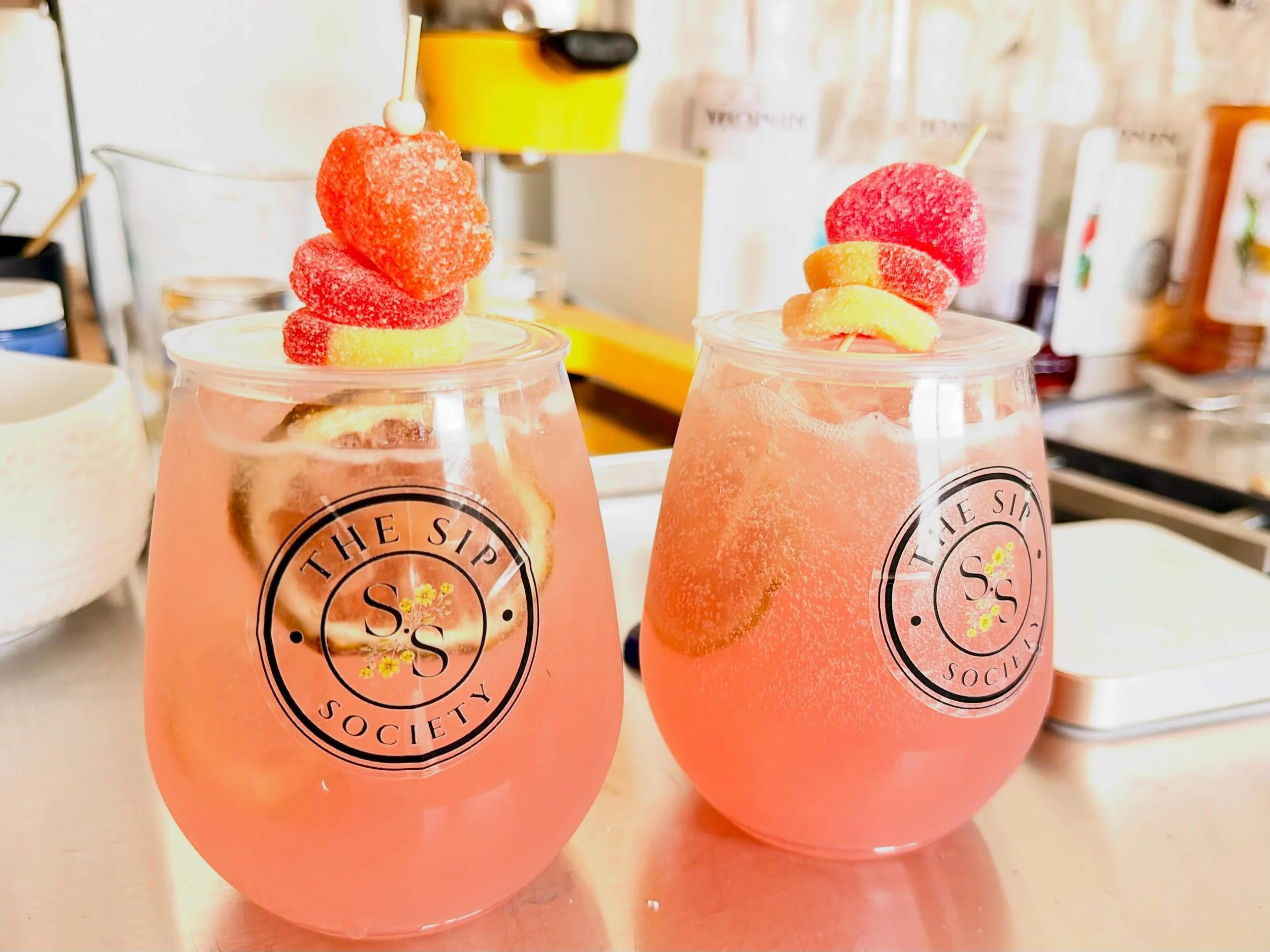 Two pink drinks in clear cups with black logos, topped with colorful gummy candies and fruit slices, on a countertop in a café or restaurant setting.