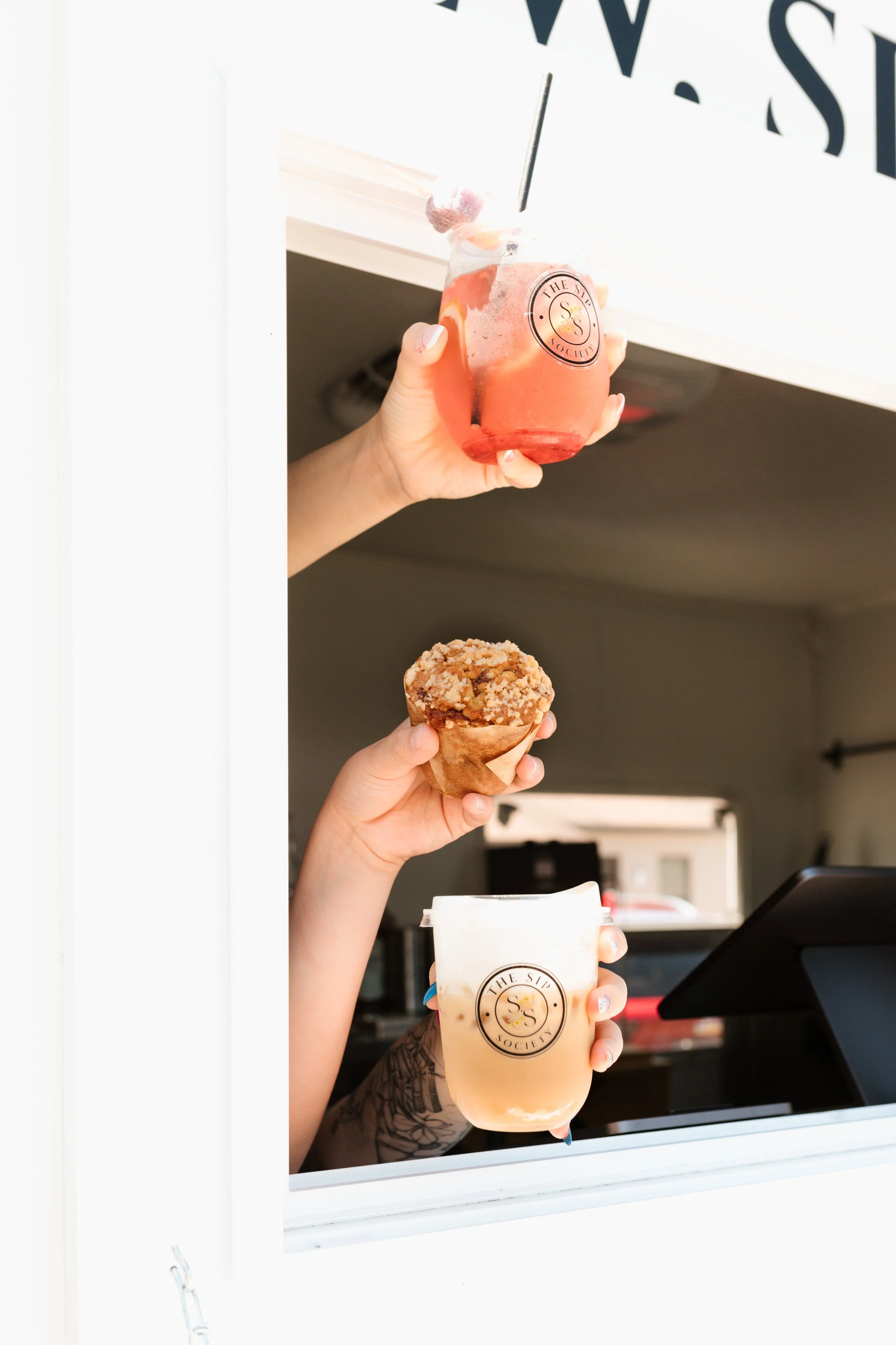 A person holding a pink drink in a clear cup with a black straw and a strawberry on top, seen through a drive-thru window. In the person's other hand, they hold a caramel apple and a large coffee cup.