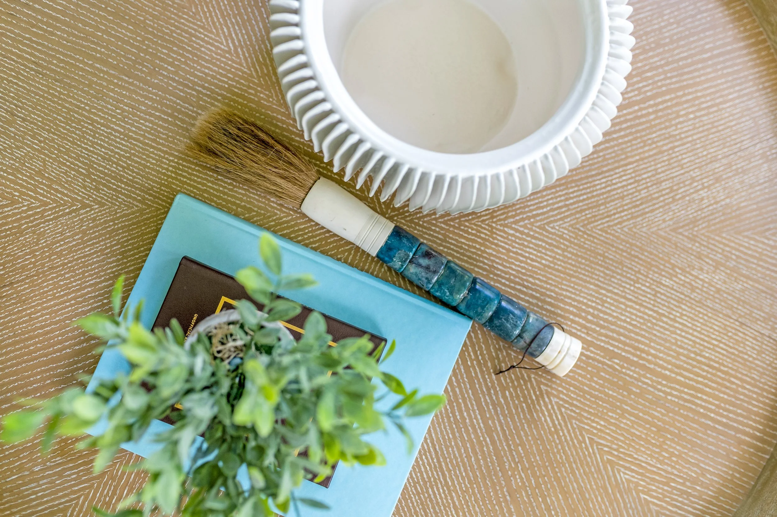 Top-down view of a small plant, a blue book with a black cover, a brush with a white handle and some bristles, a white decorative frame with a textured pattern, all placed on a beige textured surface.