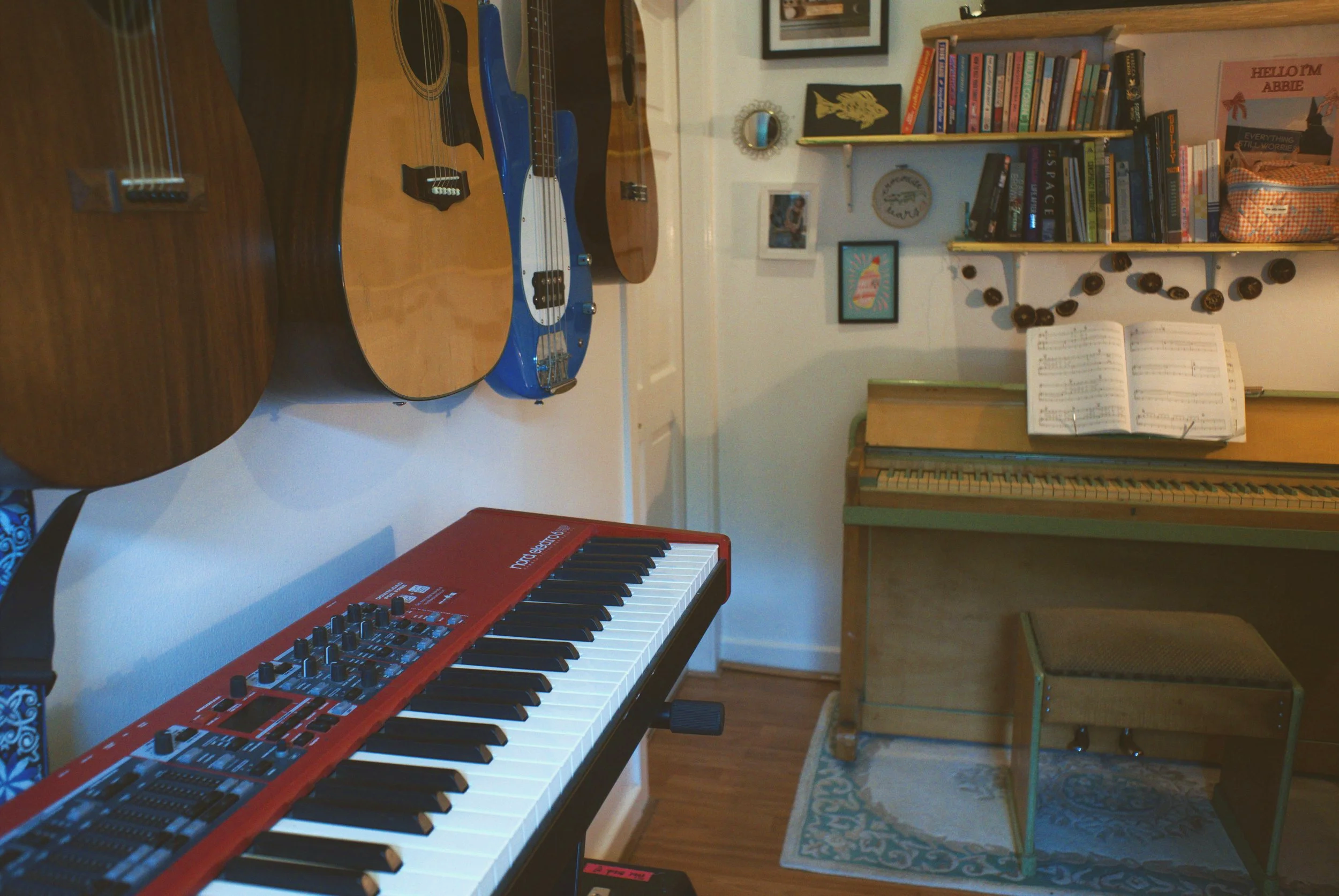 A room with musical instruments including an electric keyboard, an acoustic guitar, and a bass guitar, a piano with sheet music, and a bookshelf with books and decorations.