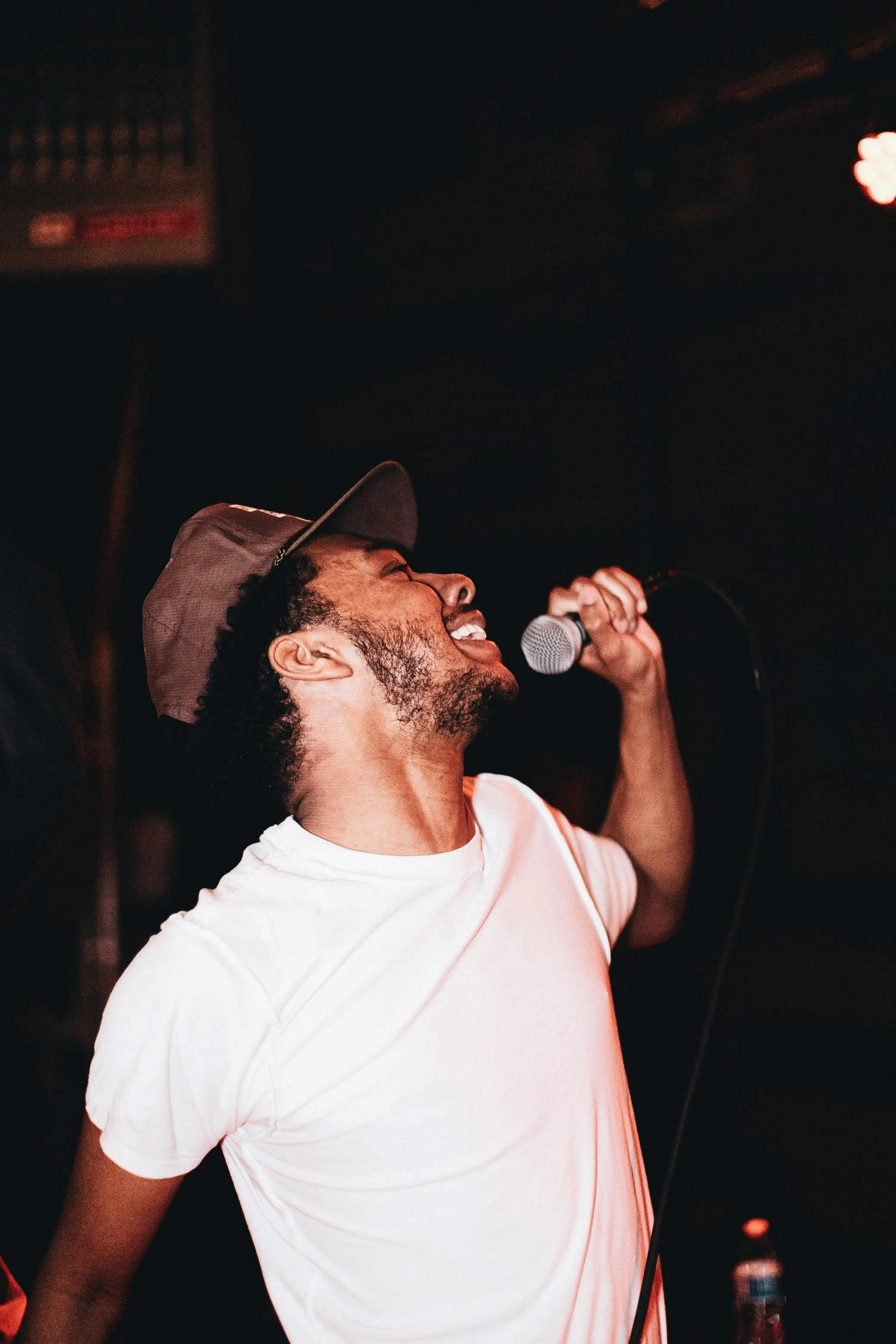 A man singing into a microphone on stage, wearing a white t-shirt and a brown baseball cap, with a joyful expression.