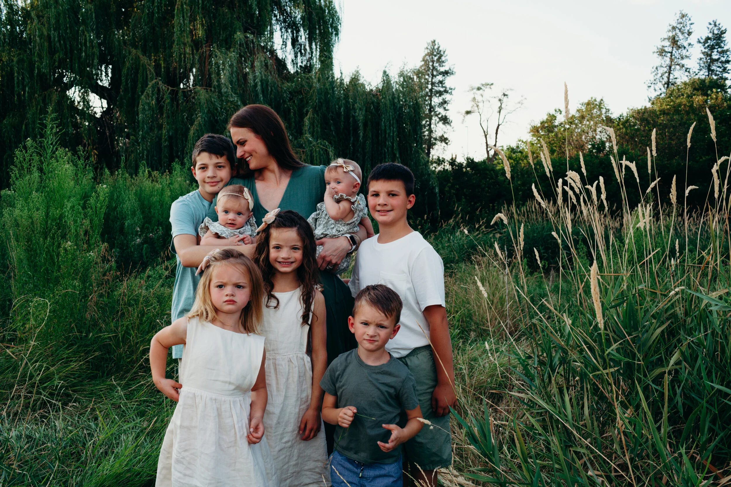 Andrea Durham with her children standing outdoors in a grassy area surrounded by trees, some children are smiling while others look serious.