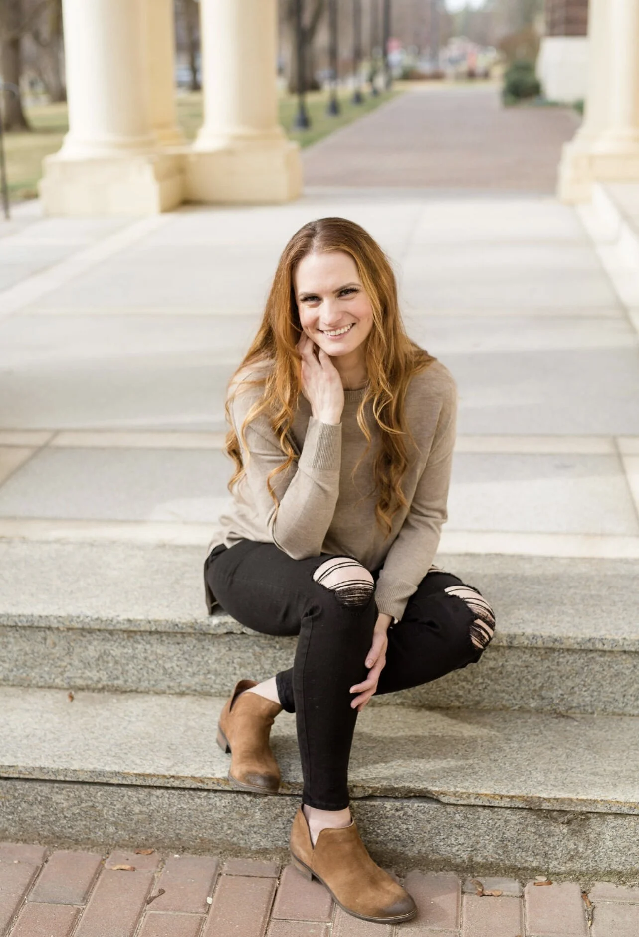 Andrea Durham, Midife and Founder of The Catching House with long red hair smiling while sitting on outdoor steps near large columns in a park.