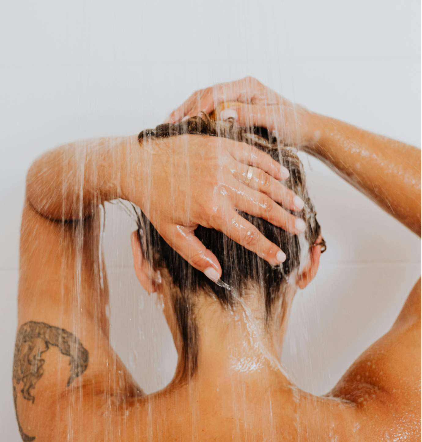 A person taking a shower with water streaming down their head and shoulders, hands on their head, showing wet hair and a tattoo on the arm.
