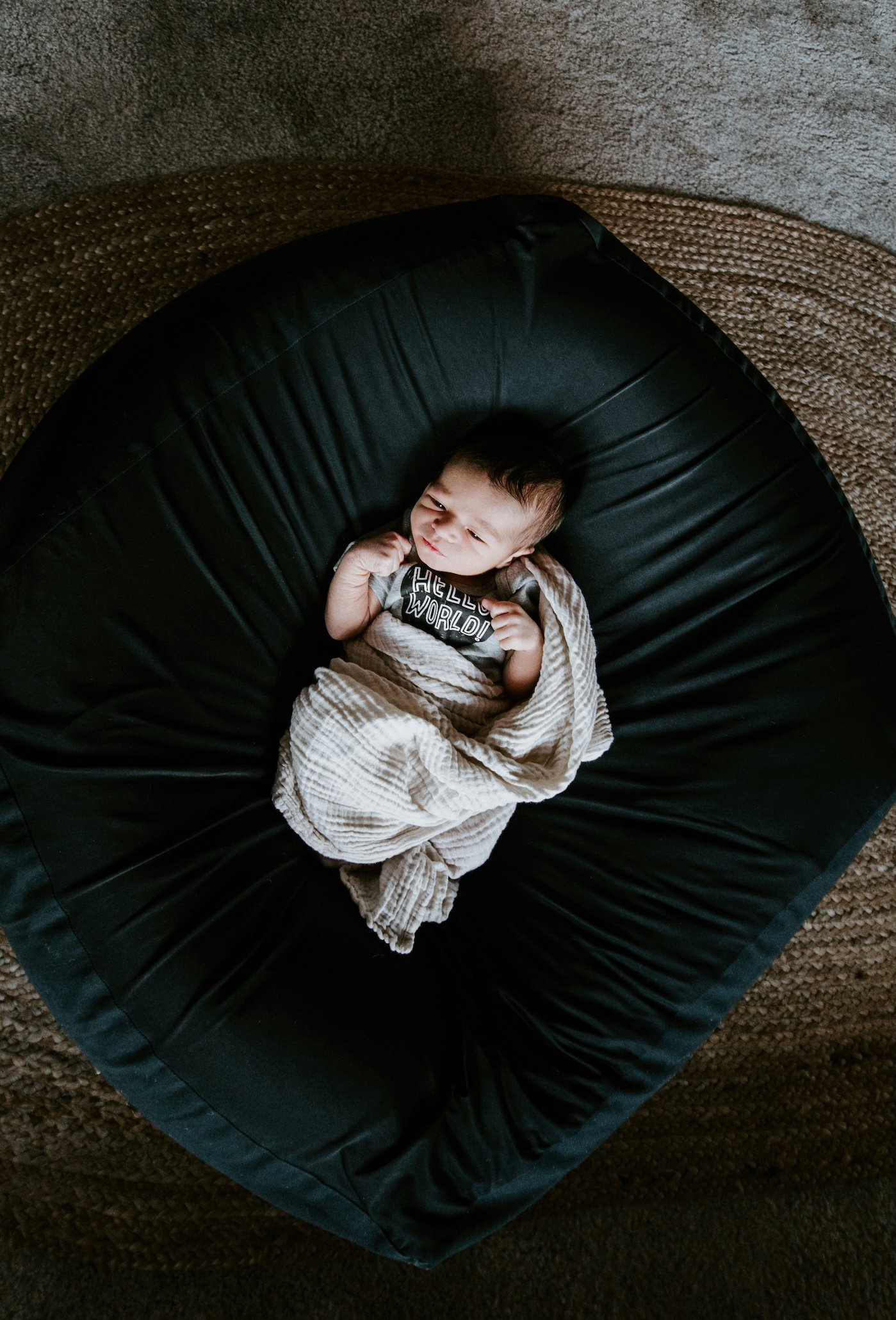 A young child lying on a black circular chair, wrapped in a beige striped blanket, wearing a T-shirt that says 'Hello World!'