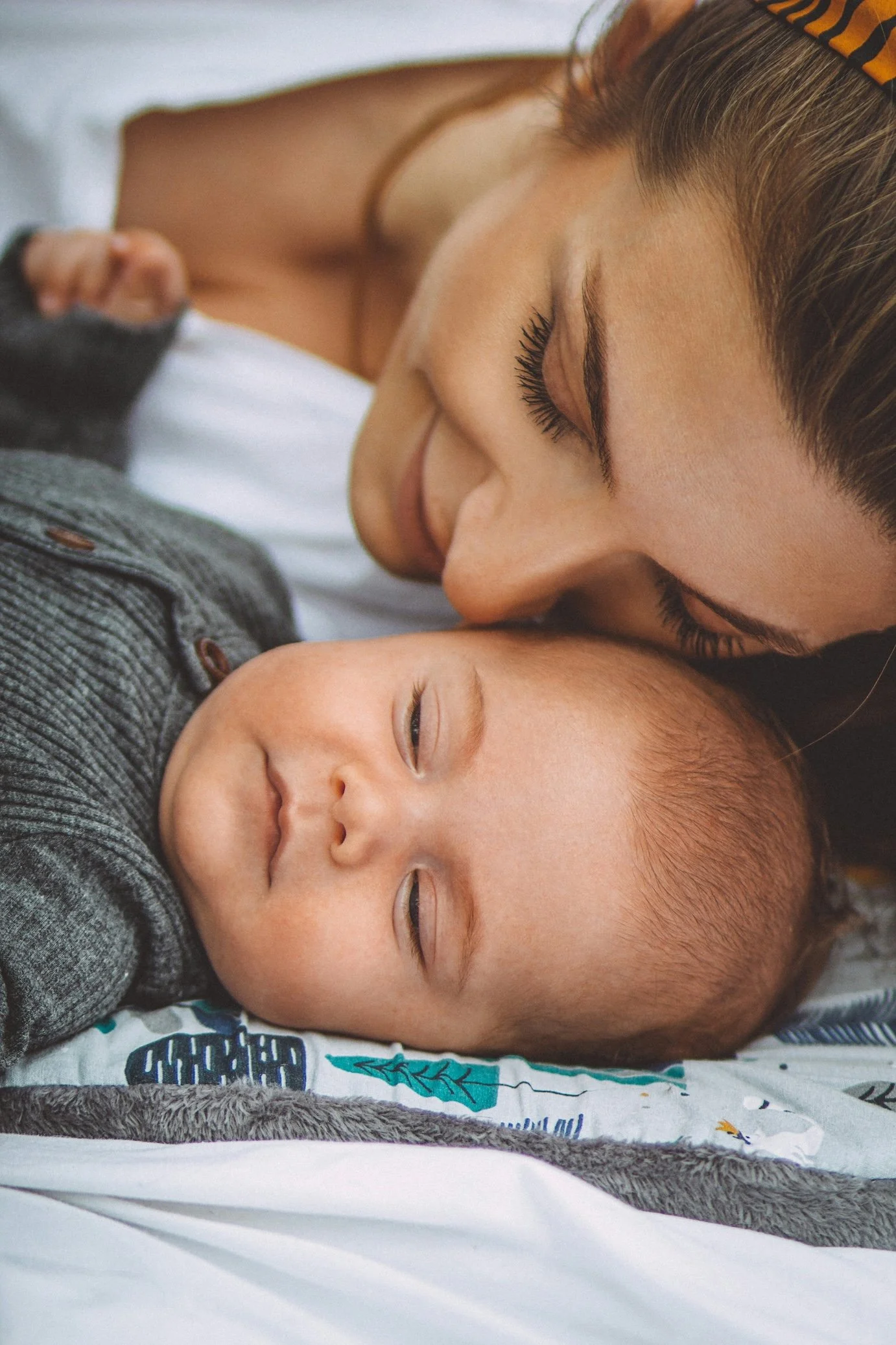 A woman with red hair and closed eyes lightly kisses a smiling baby with closed eyes resting on a bed.