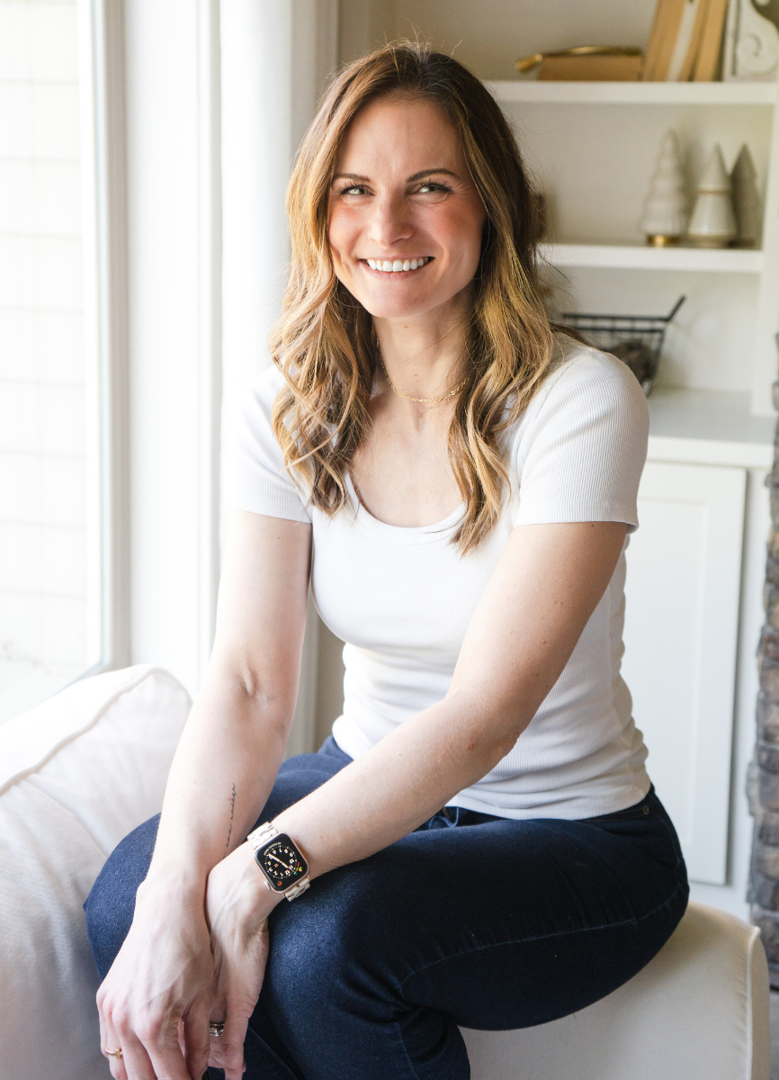Andrea Durham, Midife and Founder of The Catching House on a chair in a well-lit room, wearing a white T-shirt and dark jeans, with a smartwatch on her left wrist.