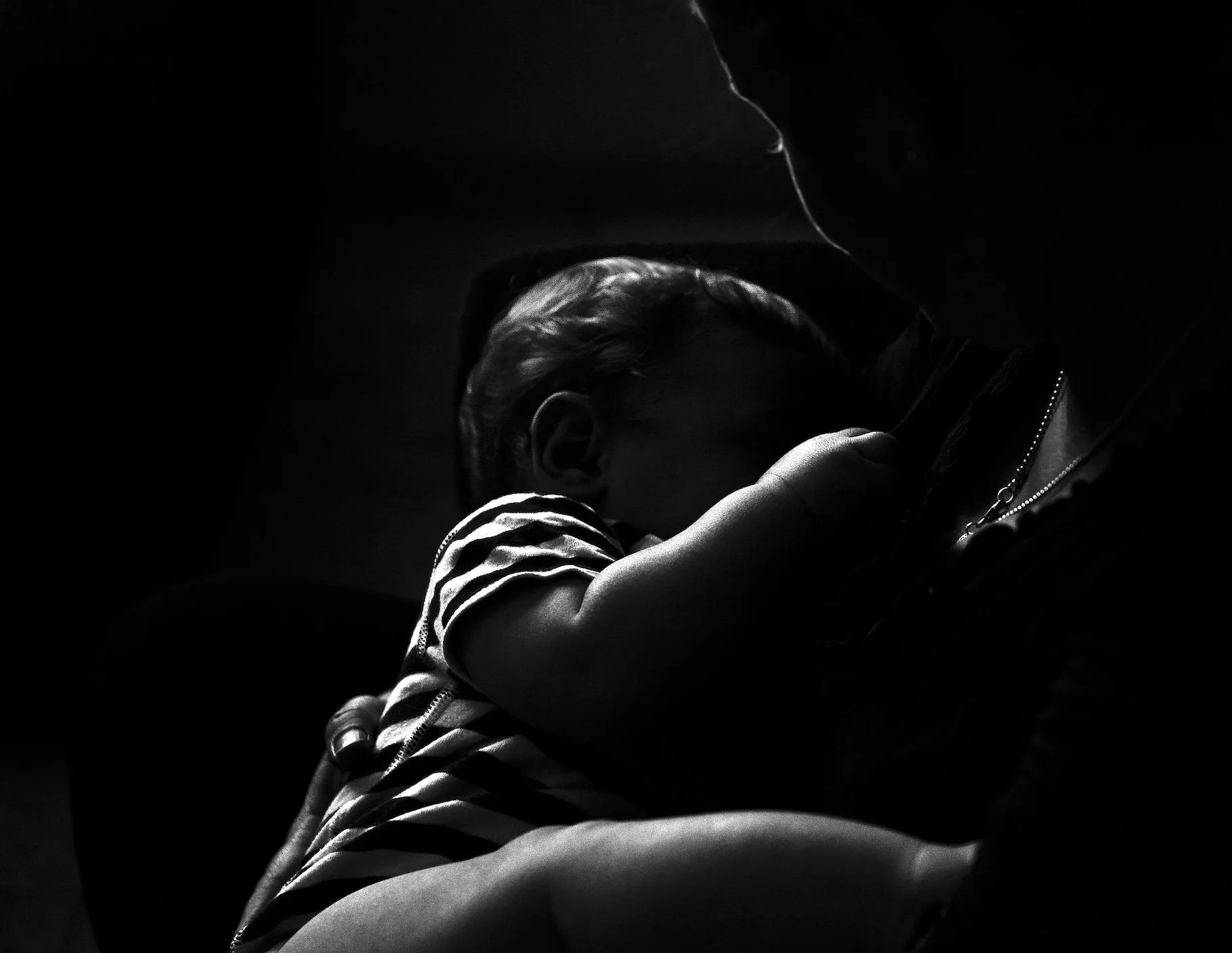A black and white photograph of an adult holding a sleeping child. The child's face is turned away, and the adult is partially visible, emphasizing the protective embrace.