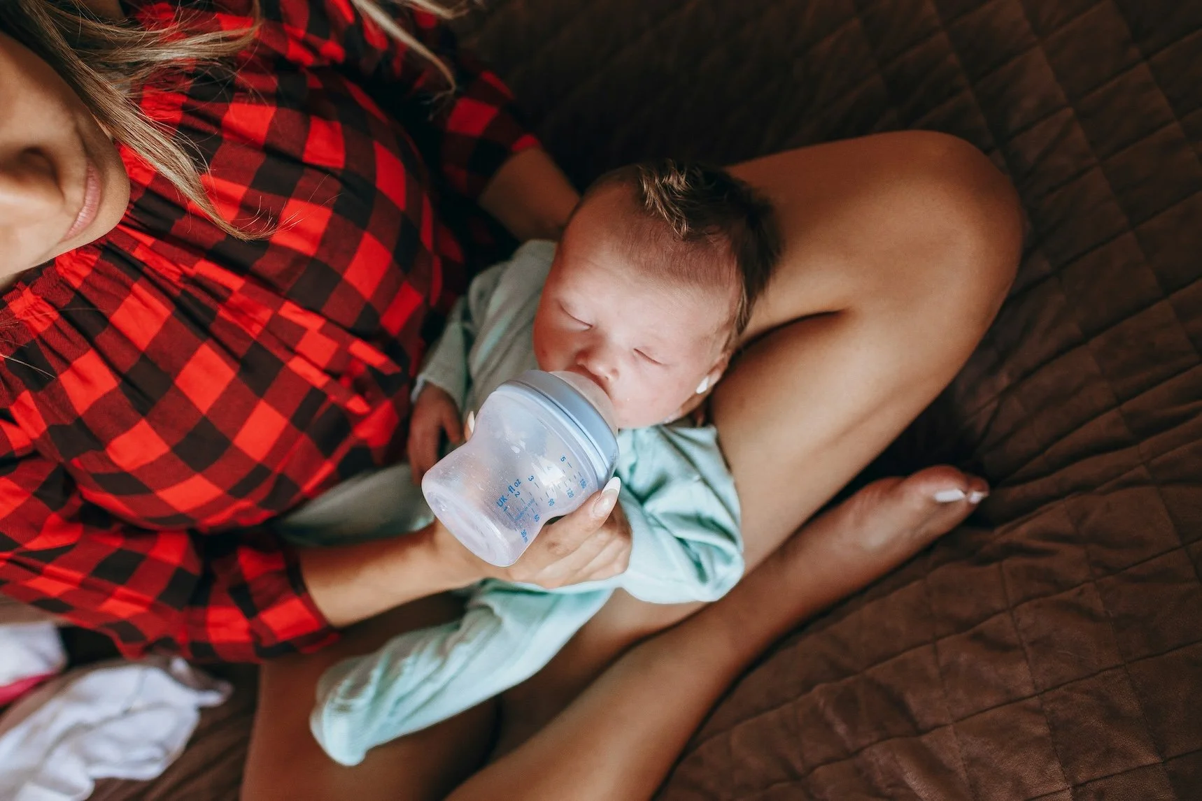 A baby lying on a woman's lap, drinking milk from a bottle. The woman is wearing a red and black checkered shirt, and the baby is dressed in a light green outfit. They are sitting on a brown textured surface.