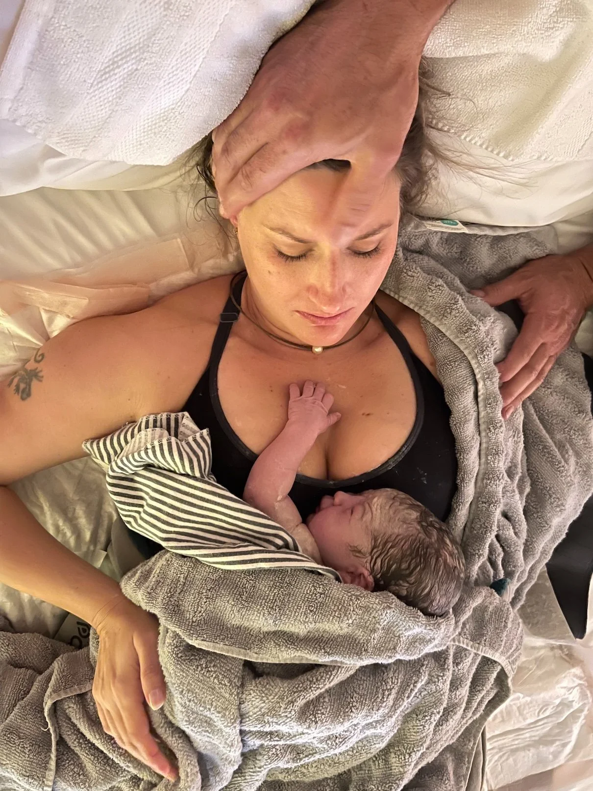A woman lying on a bed with a newborn baby on her chest, with a healthcare worker's hand supporting her head and another hand on her shoulder.