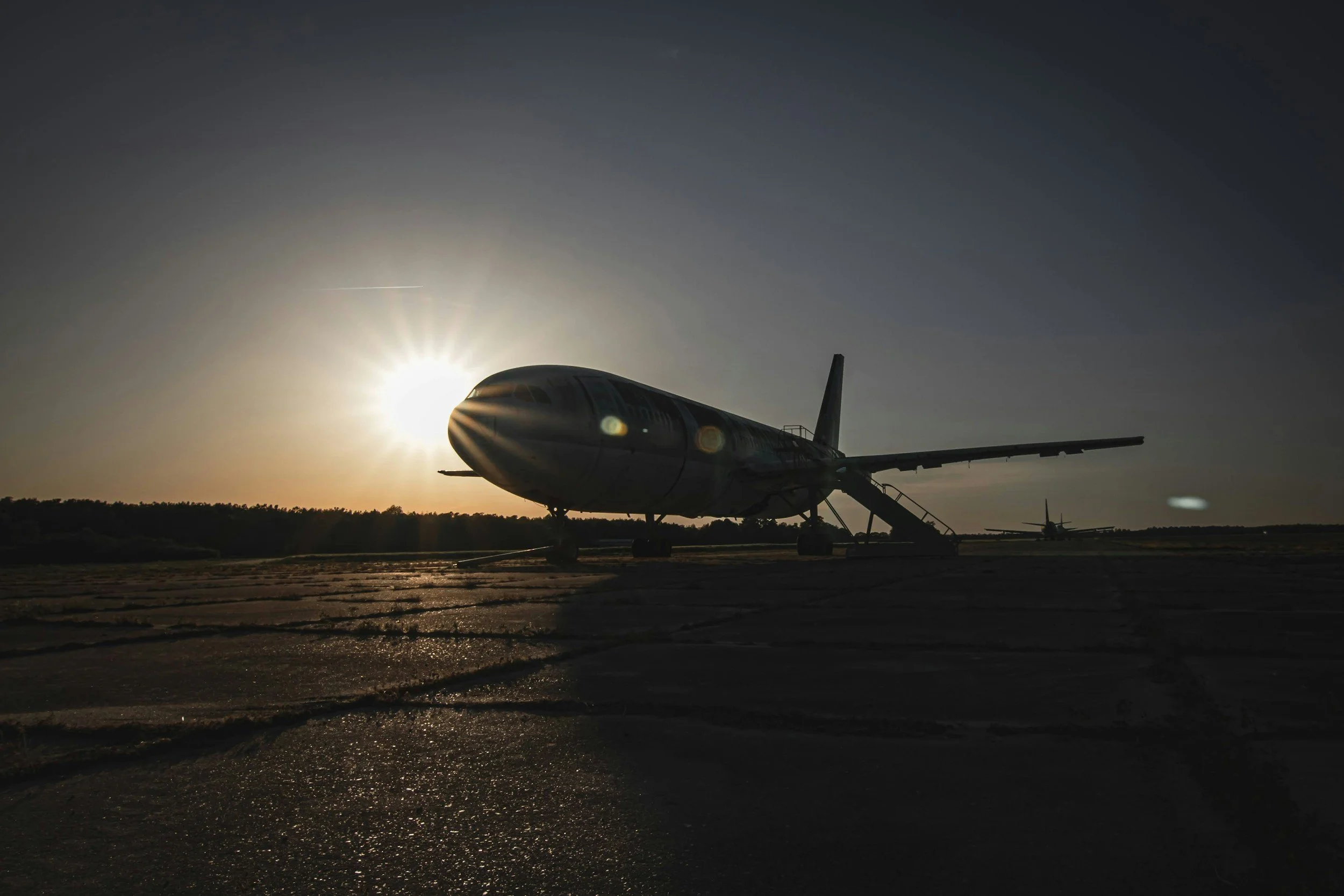 An airplane parked on a runway during sunset, with the sun casting long shadows and lens flare visible.
