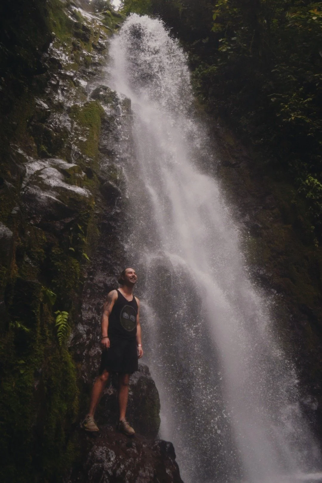 A man standing on rocks in front of a waterfall in a forest, smiling and looking up at the water.