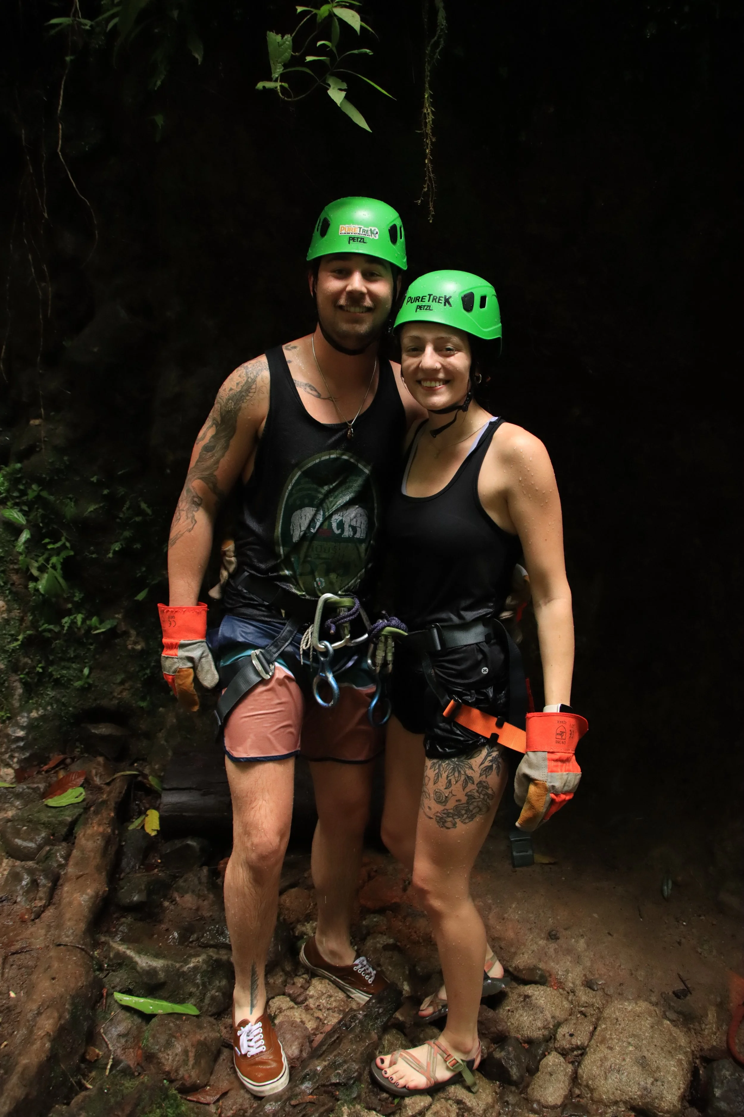 A man and woman in black tank tops and helmets standing on wet rocks in a jungle, smiling, with lush green plants around them.