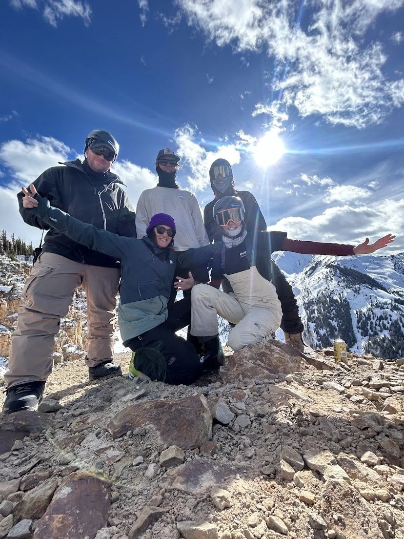 Five people dressed in winter gear posing on a rocky mountain with snow-covered peaks in the background and a bright sun shining through partly cloudy sky.