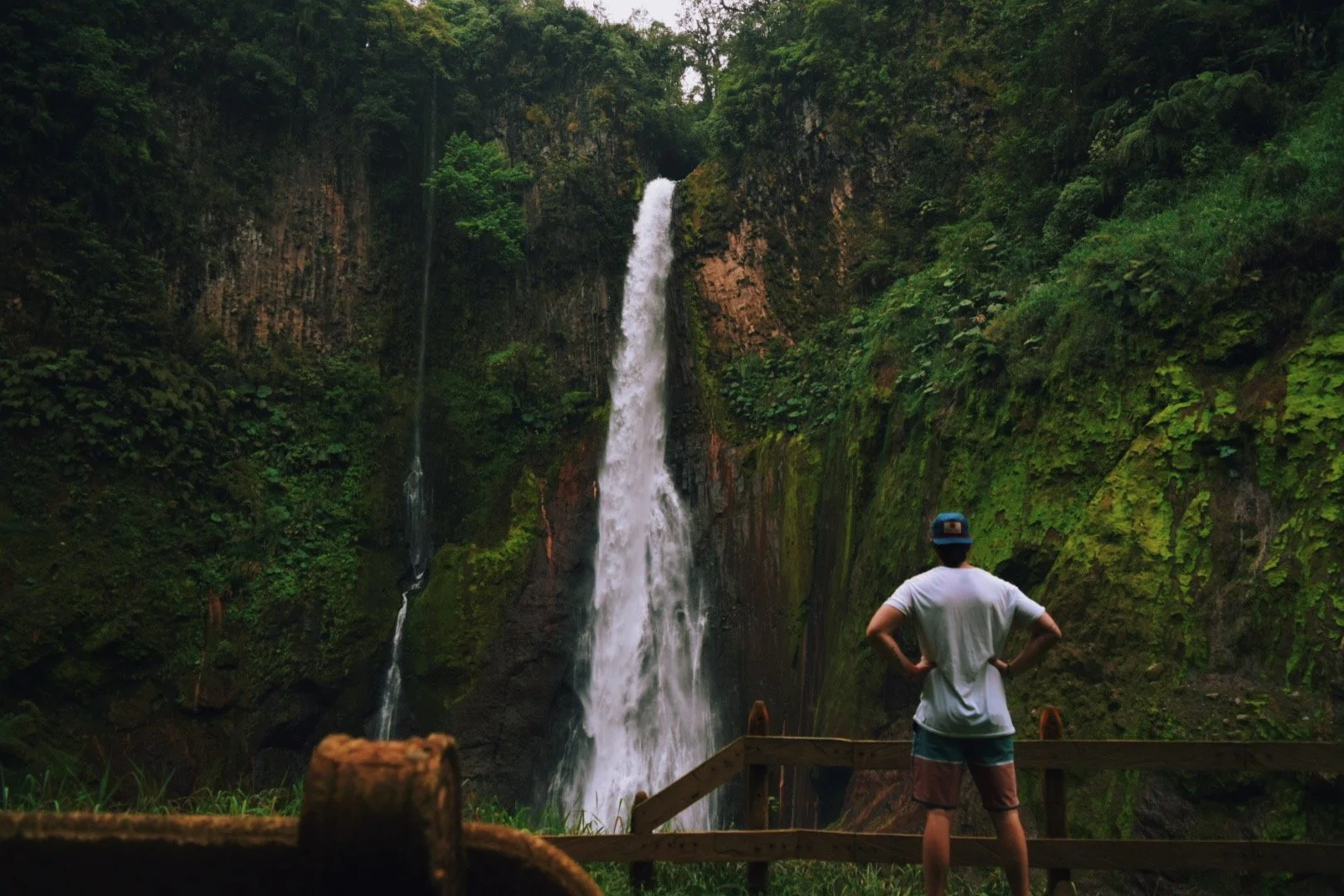 A person standing on a wooden viewing platform with hands on hips, looking at a tall waterfall surrounded by lush green forest.
