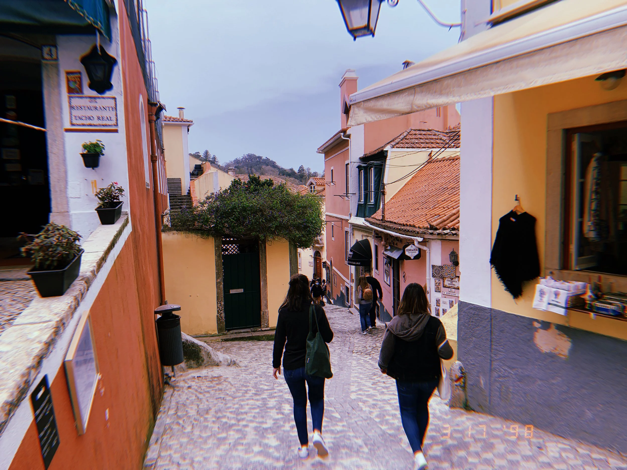 People walking down a cobblestone street with colorful buildings and shops, some with awnings and hanging signs, on a cloudy day.