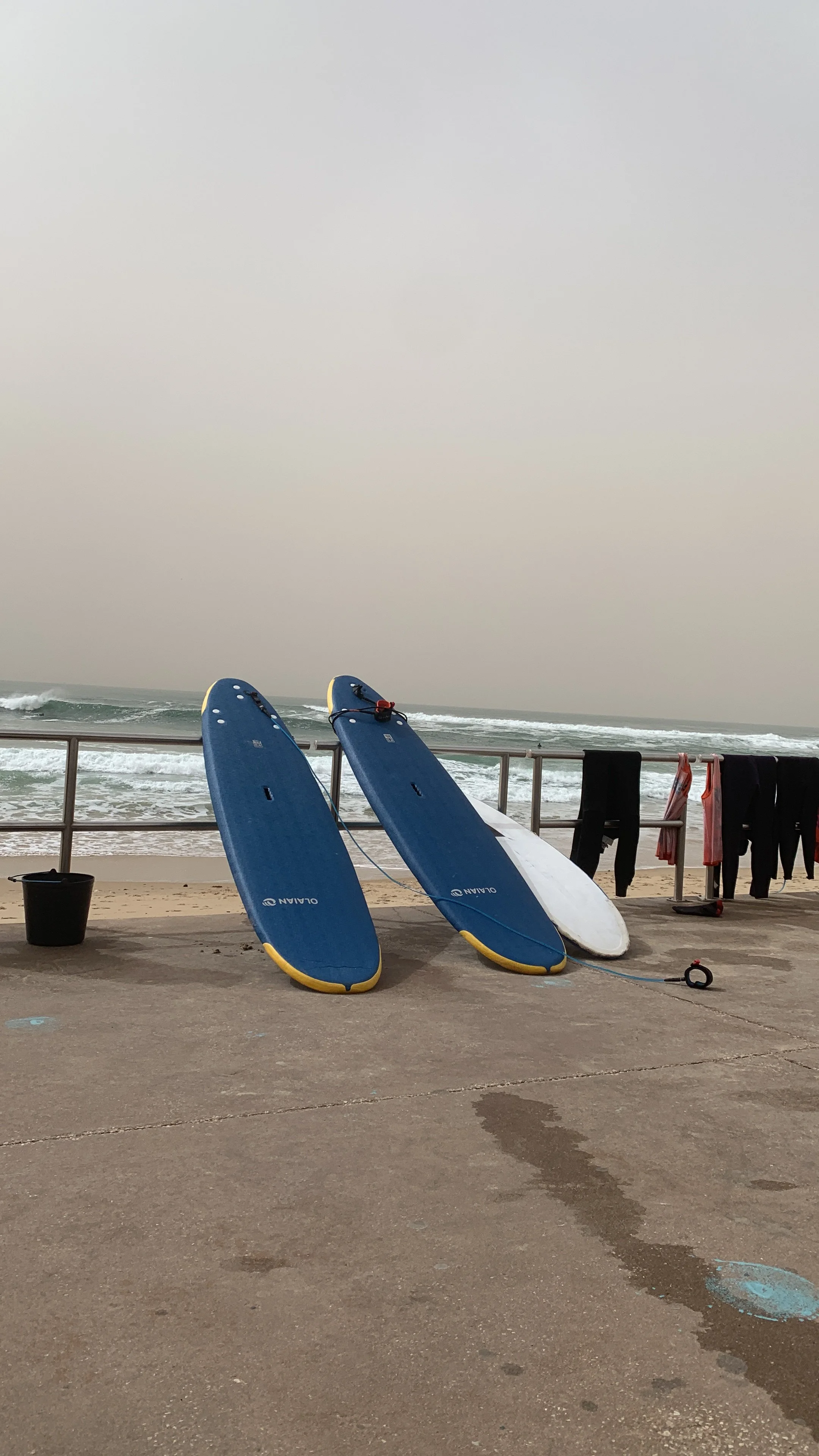 Two blue surfboards resting against a small fence on a beach, with ocean waves in the background and clothes hanging to dry on a line.