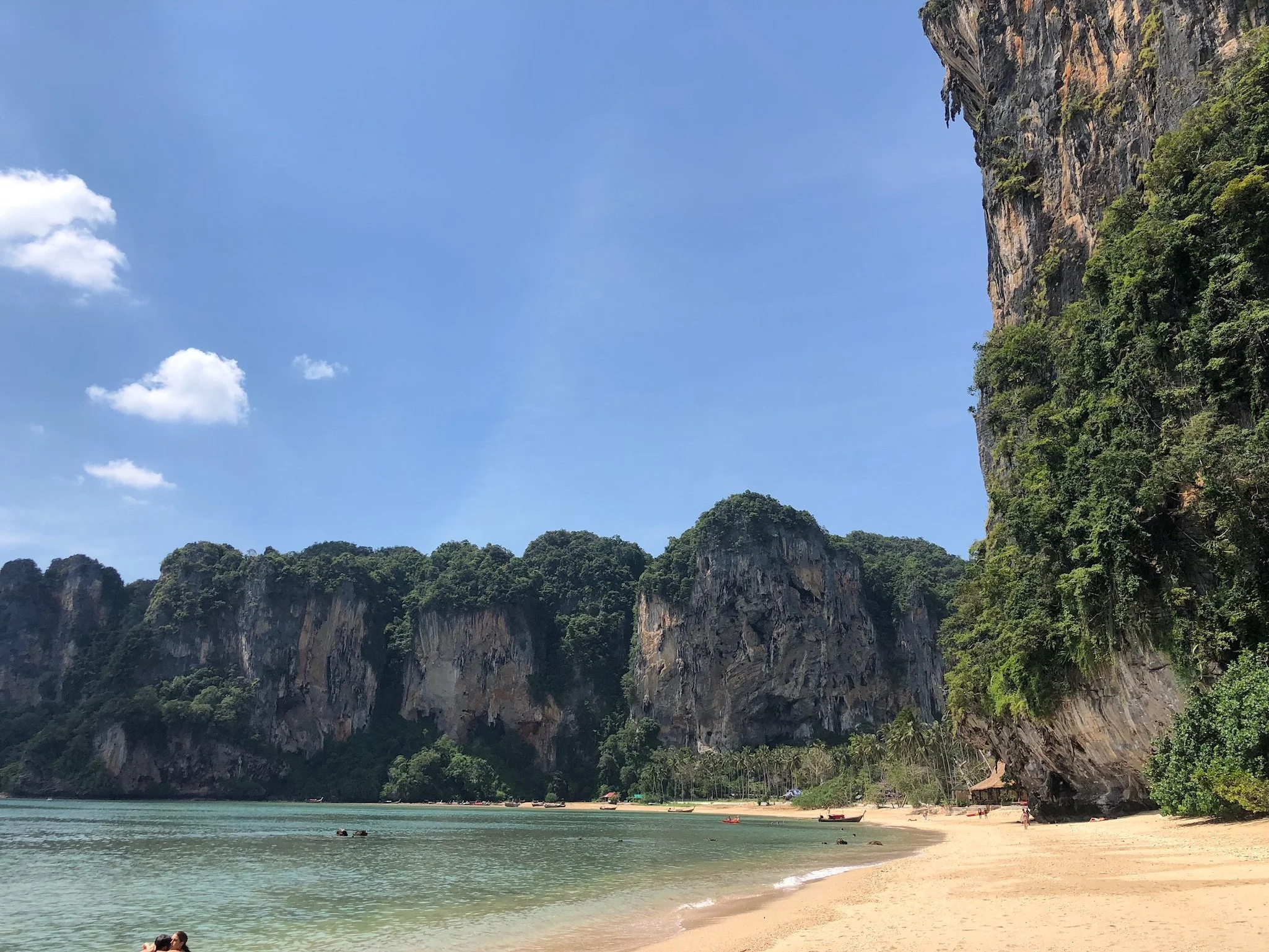 Beach with clear water, small boats, and a backdrop of lush green mountains and cliffs under a blue sky with a few clouds.