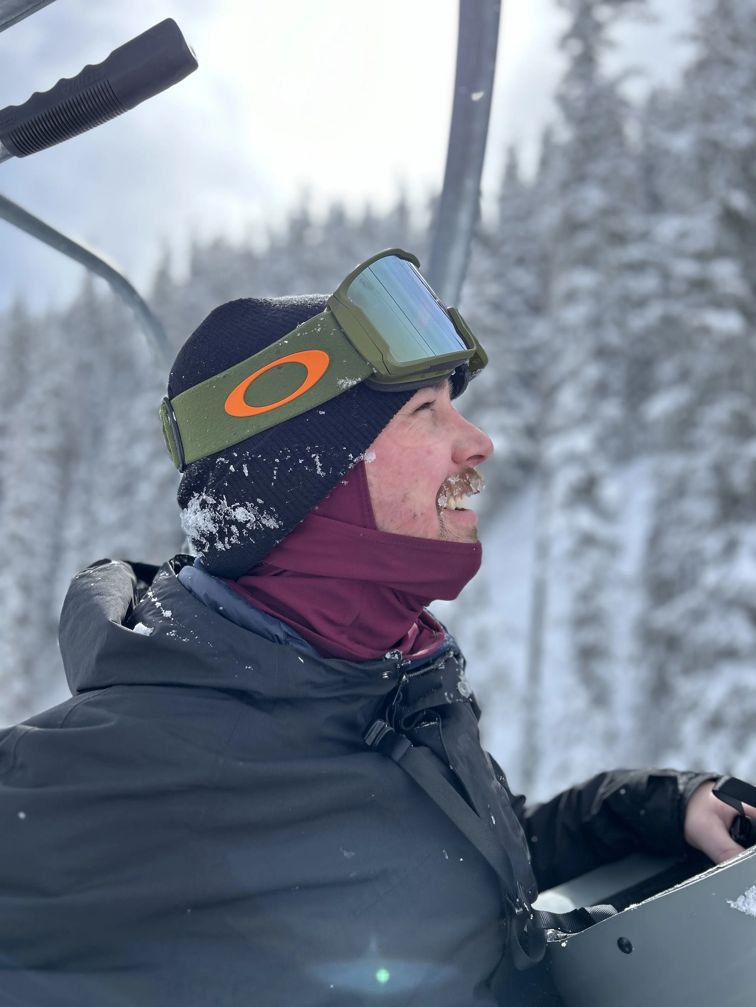 A person with a beard, wearing dark ski clothes and green goggles, riding a chairlift in a snowy forest with trees and a blue sky in the background.