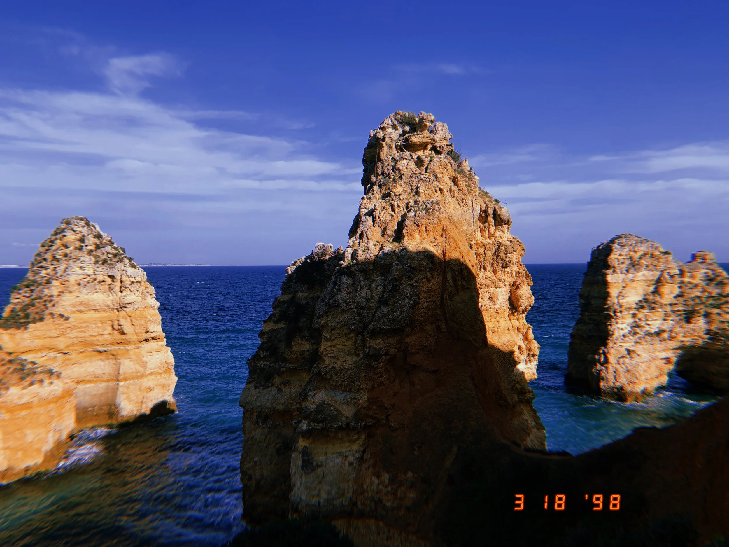 Three large rock formations jut out of the ocean against a blue sky with scattered clouds.