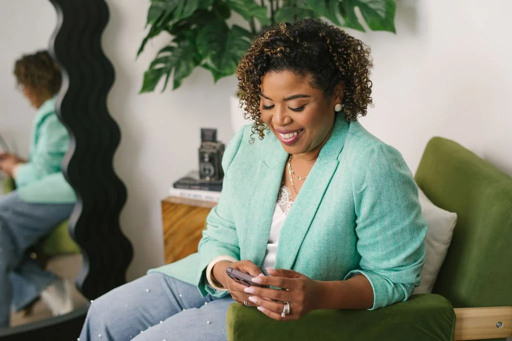Smiling woman with curly hair sitting on a green couch, looking at her phone, with a woman in the background sitting in a chair and texting, in a room with a large plant and a wavy mirror.