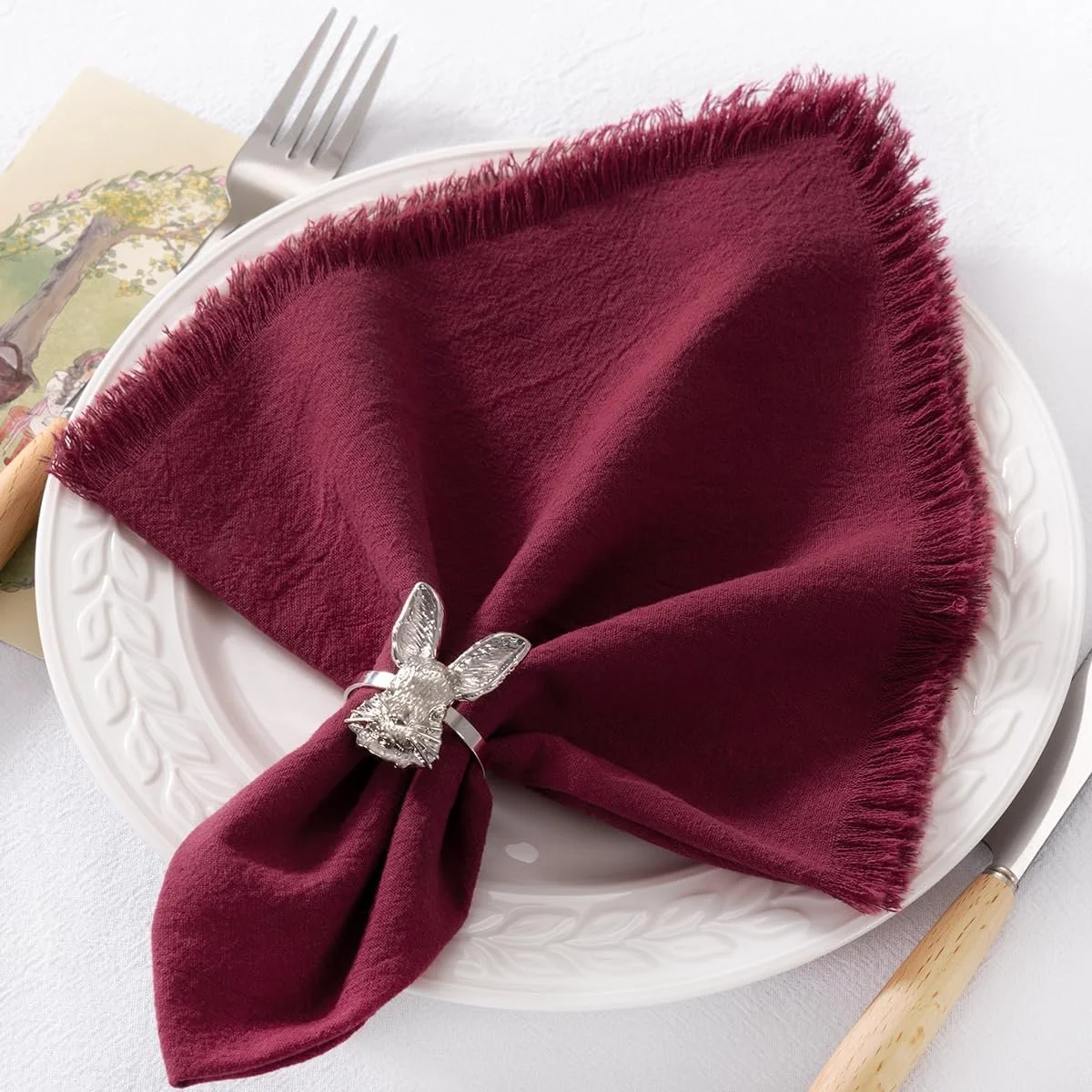 A white plate with a burgundy cloth napkin folded on it, secured with a silver bunny-shaped napkin ring, set on a white table with a fork and knife nearby.