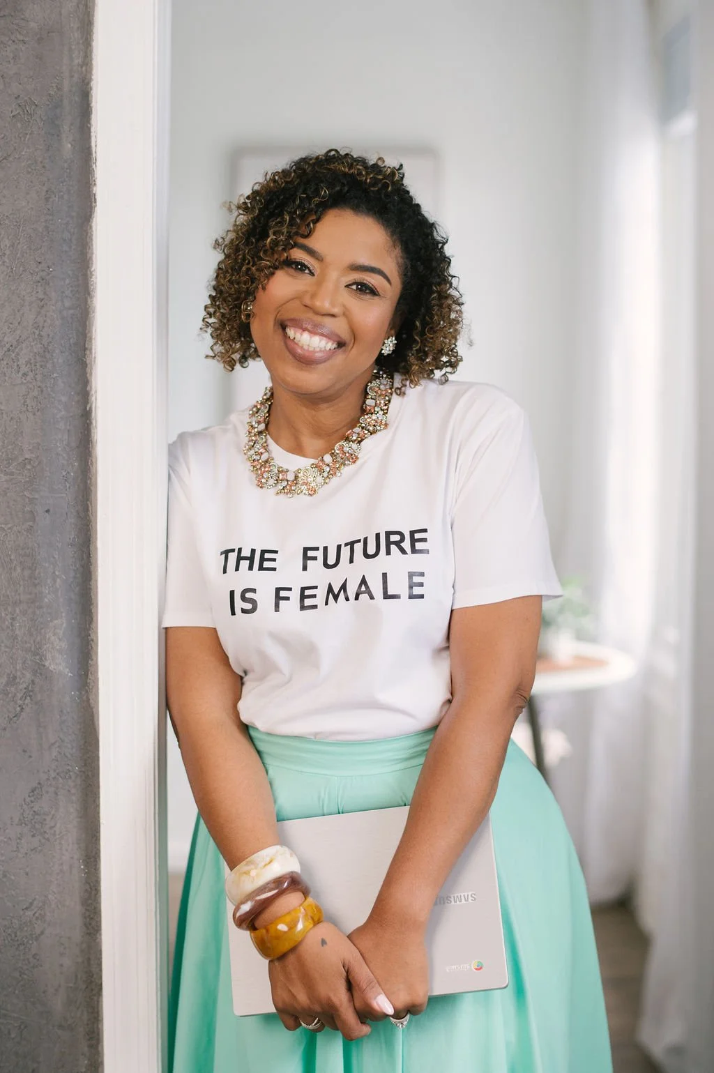 A woman with curly hair, smiling, wearing a white t-shirt with the text 'The Future is Female,' a mint green skirt, and accessorized with multiple bangles and jewelry, standing in a bright indoor setting.