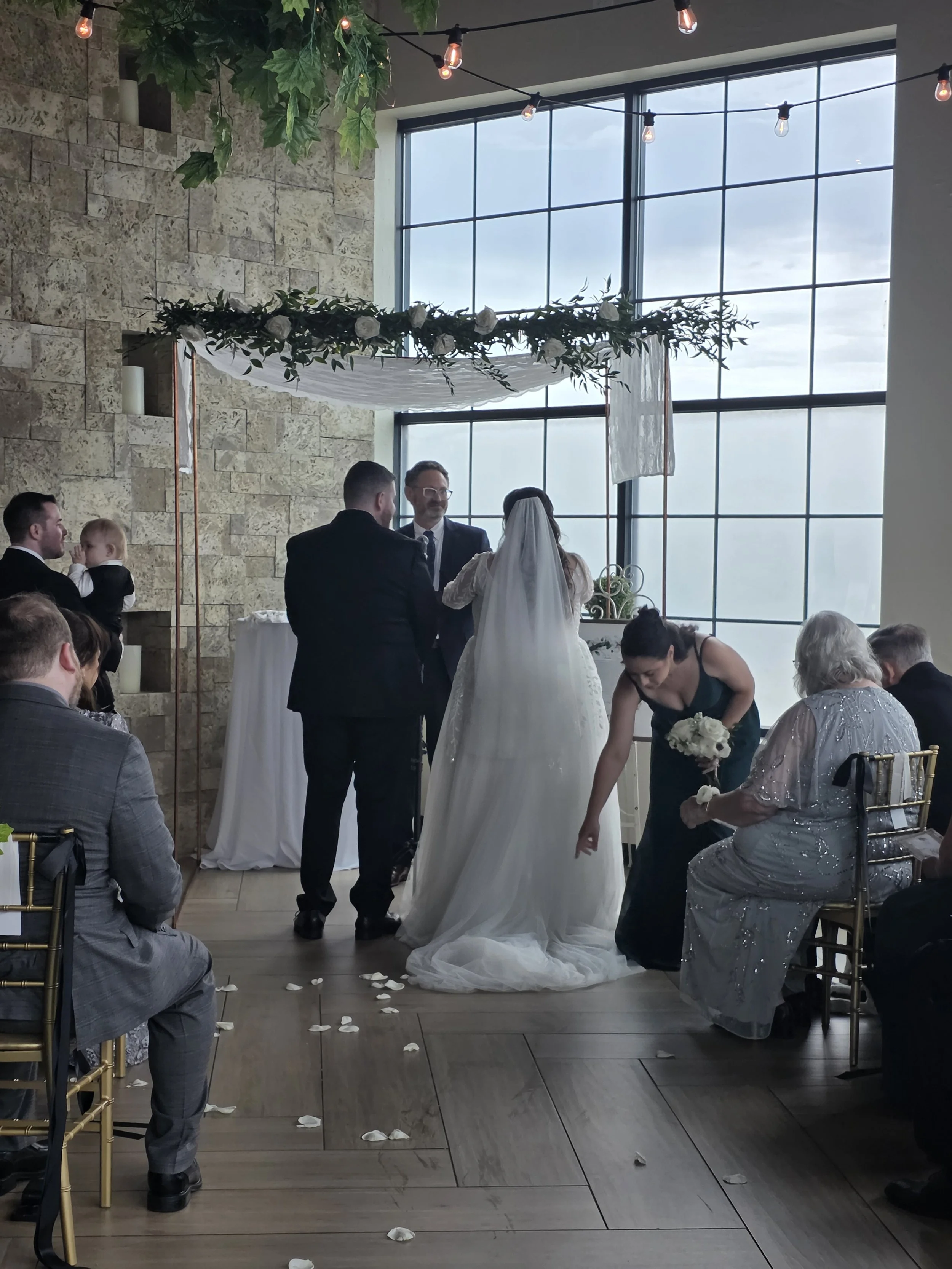 A couple getting married during a wedding ceremony in an indoor venue with large windows. The bride is wearing a white veil and dress, and the groom is in a dark suit. Guests are sitting in chairs, and flower petals are scattered on the wooden floor.