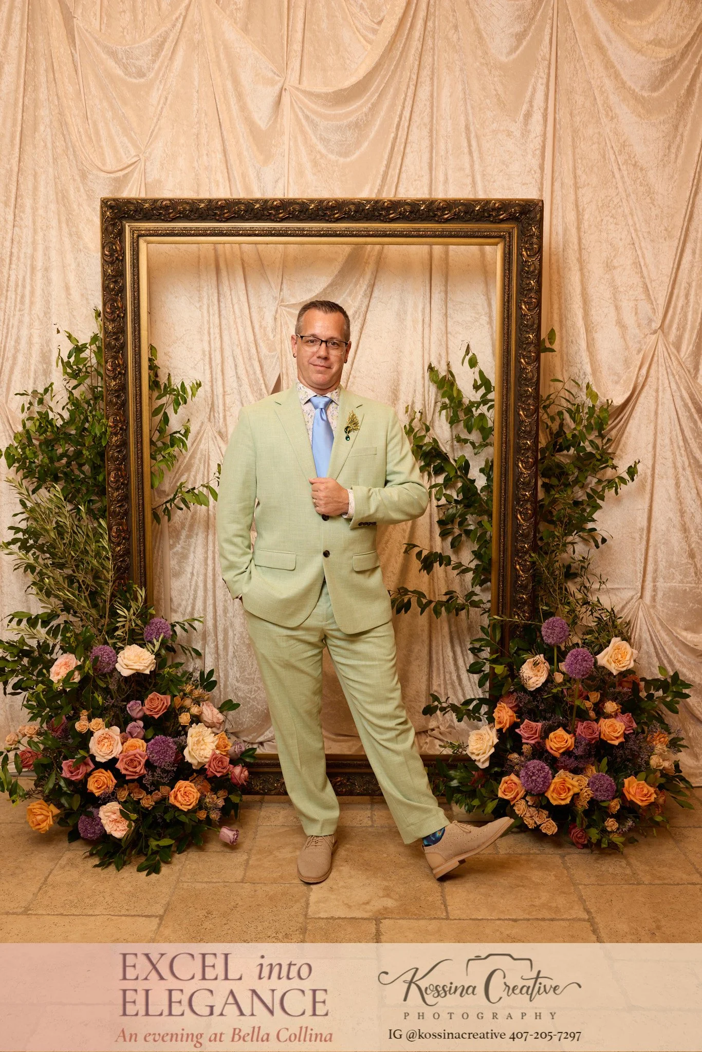 A man dressed in a light green suit standing in front of a beige curtain backdrop, surrounded by flowers. A frame is positioned behind him, creating a unique photo setup.