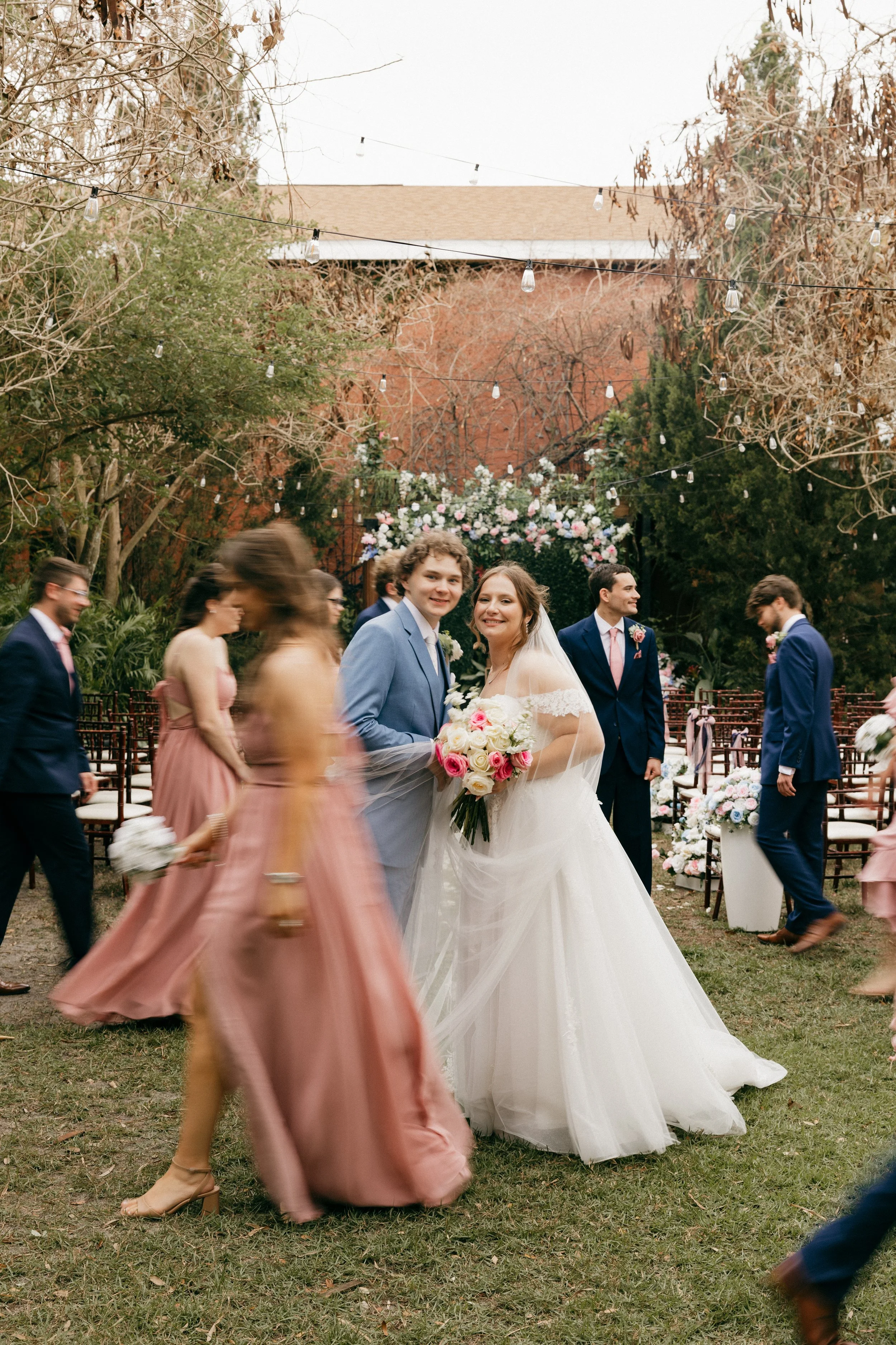 A wedding ceremony outdoors with a bride and groom in focus, surrounded by guests in formal attire, some walking or moving around. Decorations include flowers and string lights hanging overhead.