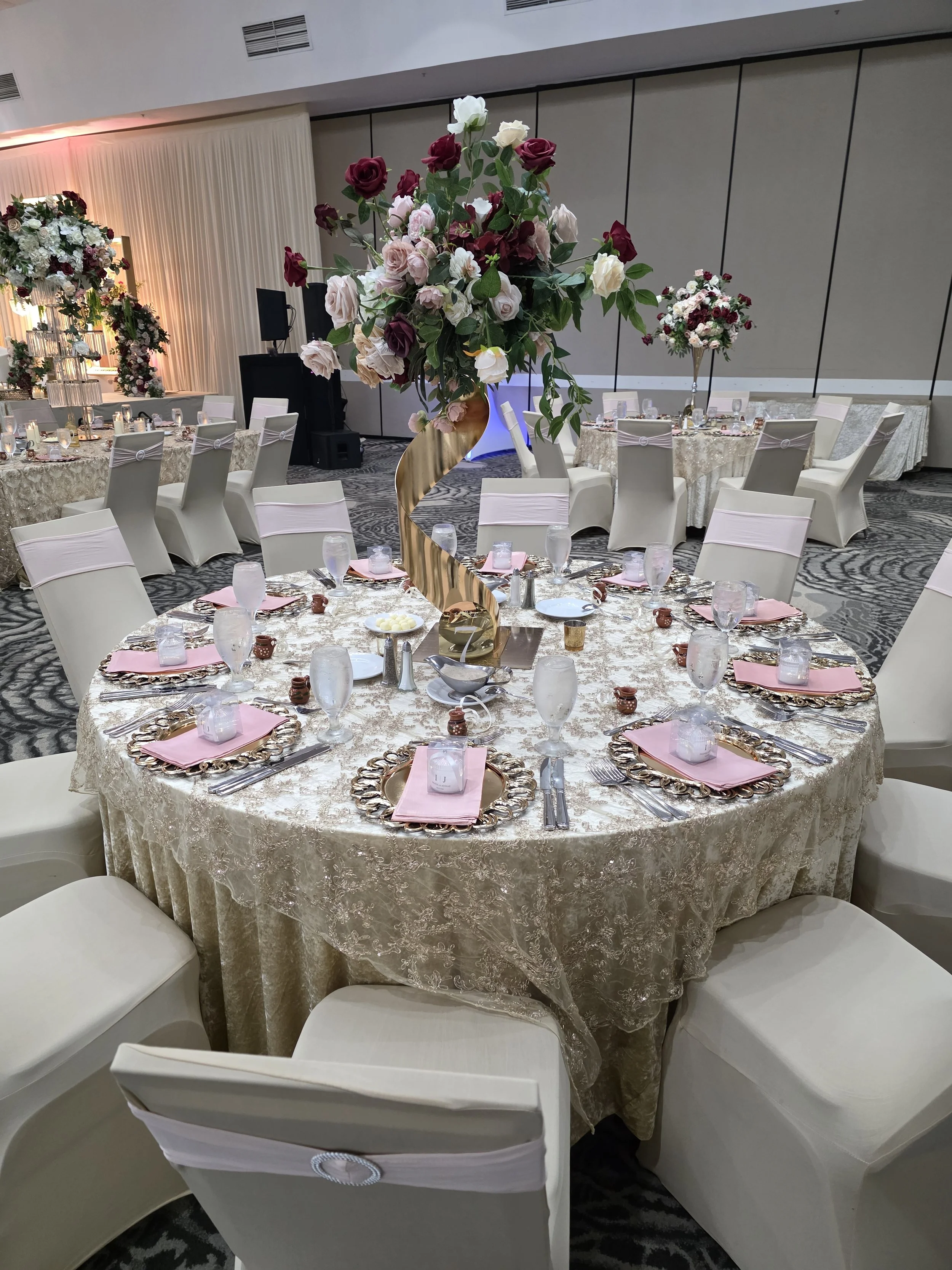 Round banquet table with a gold ornate charger plate, pink napkin, and glassware, surrounded by white chairs, with a tall floral centerpiece of pink, white, and burgundy roses, in a ballroom setting.