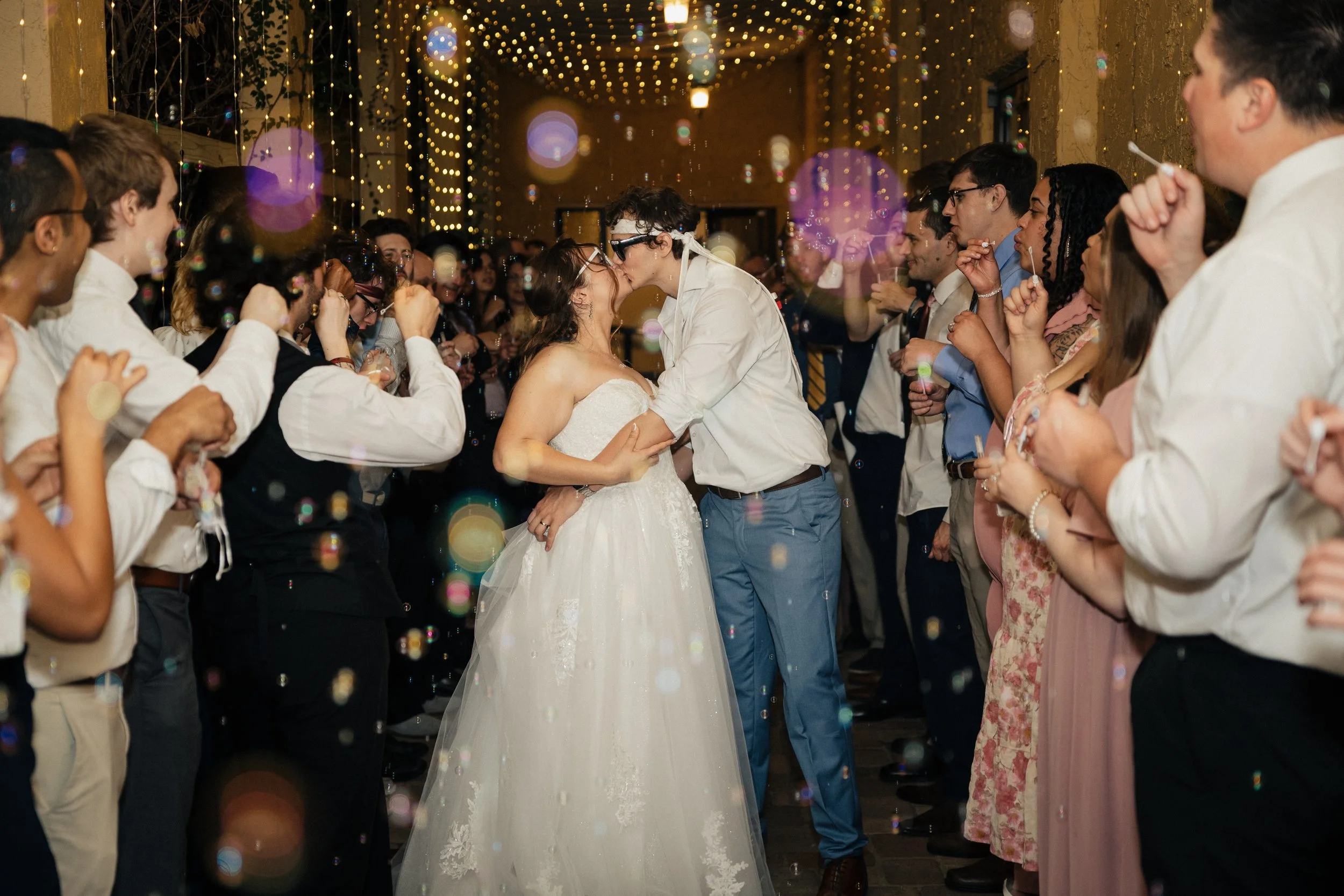 A bride and groom sharing a kiss at their wedding reception, surrounded by guests on both sides blowing bubbles, with string lights hanging overhead in a festive, decorated venue.