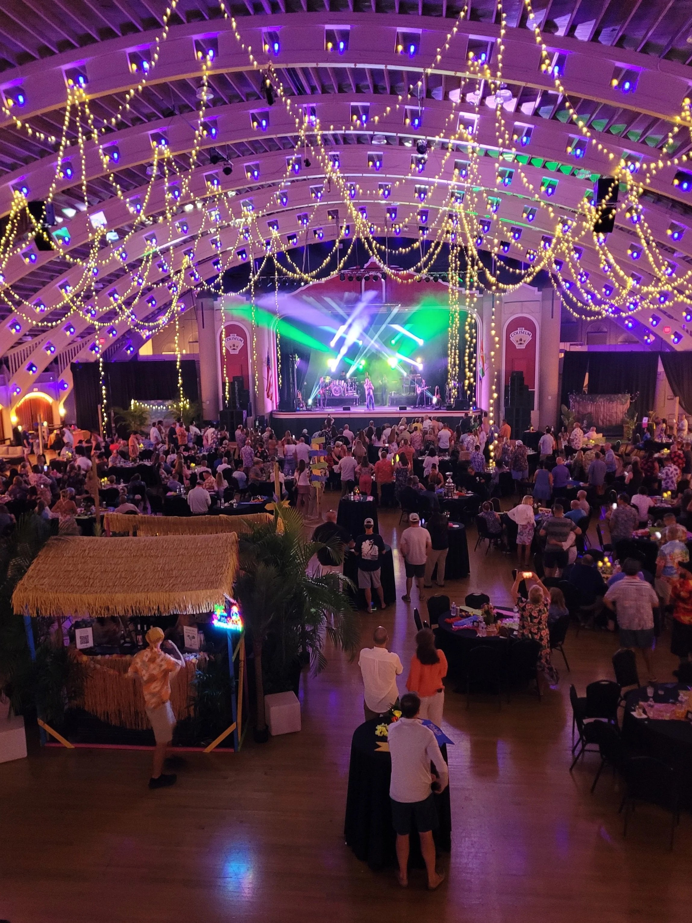 The coliseum in St. Pete set with a stage, colorful lighting, and people gathered around tables and standing. The ceiling has decorative string lights, and there is a small tiki hut-style booth in the foreground.