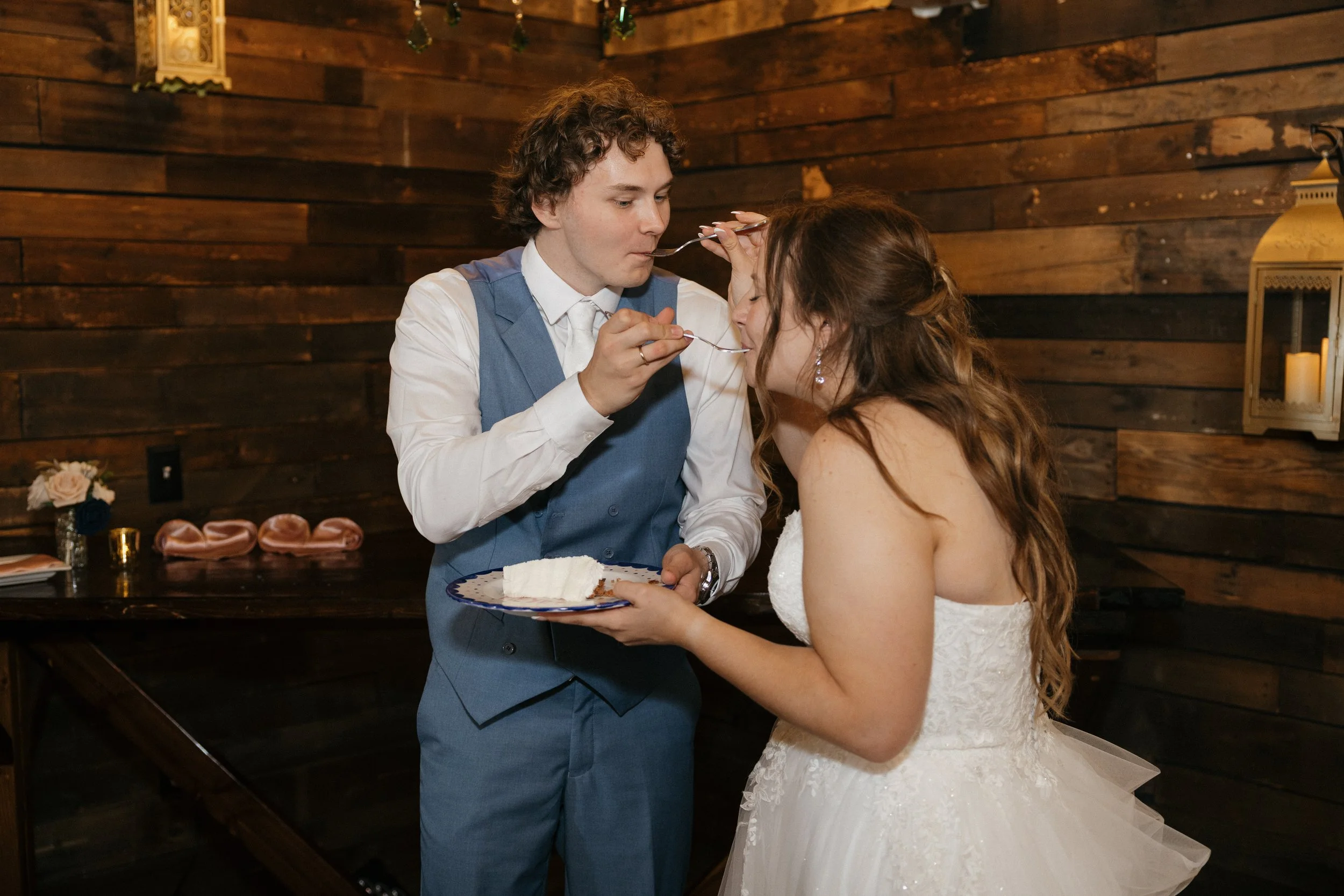 A groom feeding cake to a bride during a wedding reception in a warm, rustic decorated indoor venue with wood-paneled walls, candles, and floral arrangements.