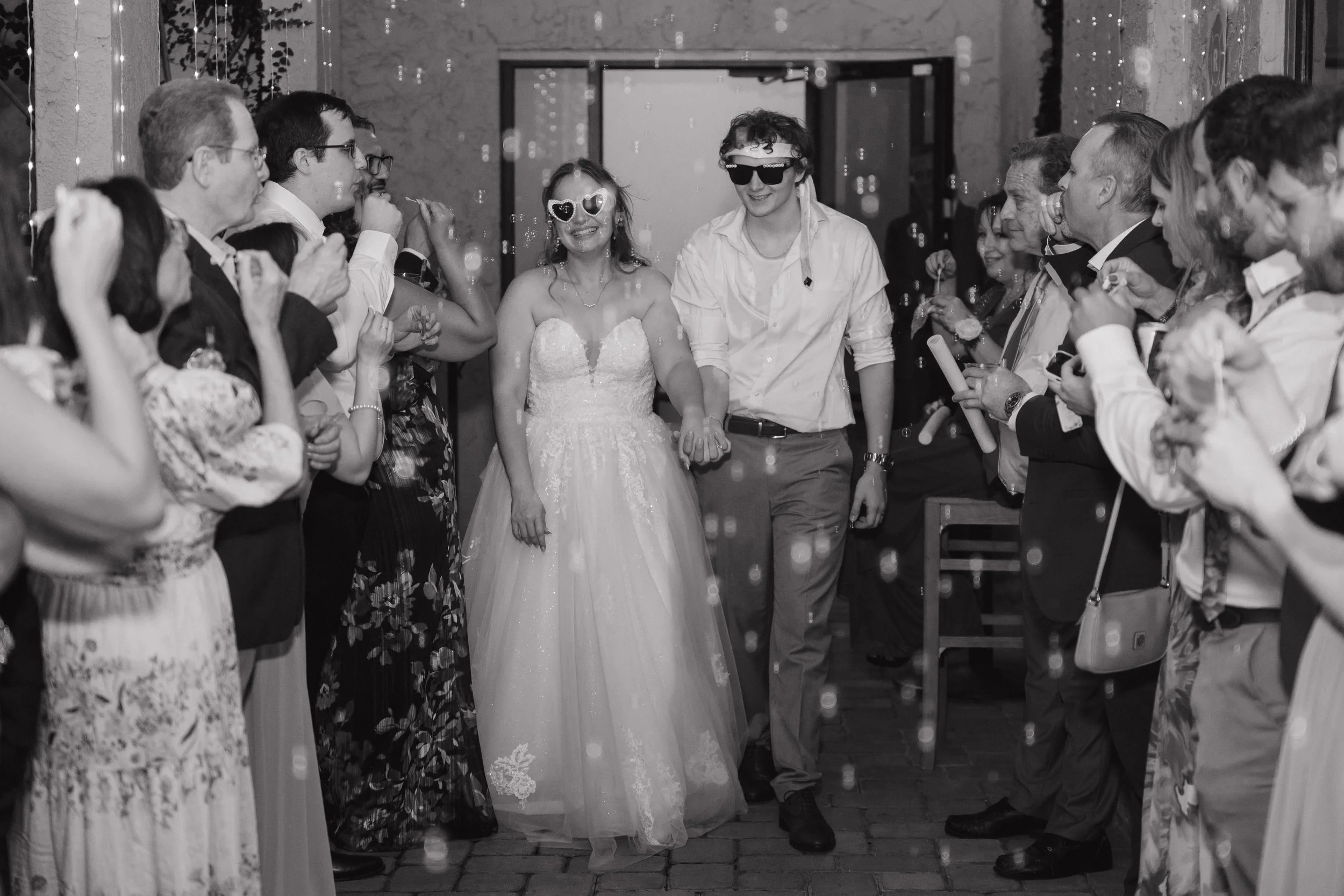 A wedding celebration with a couple holding hands amidst guests. The bride is wearing a white wedding dress with heart-shaped sunglasses, and the groom is wearing a white shirt and sunglasses. Guests are celebrating with bubbles and party favors.