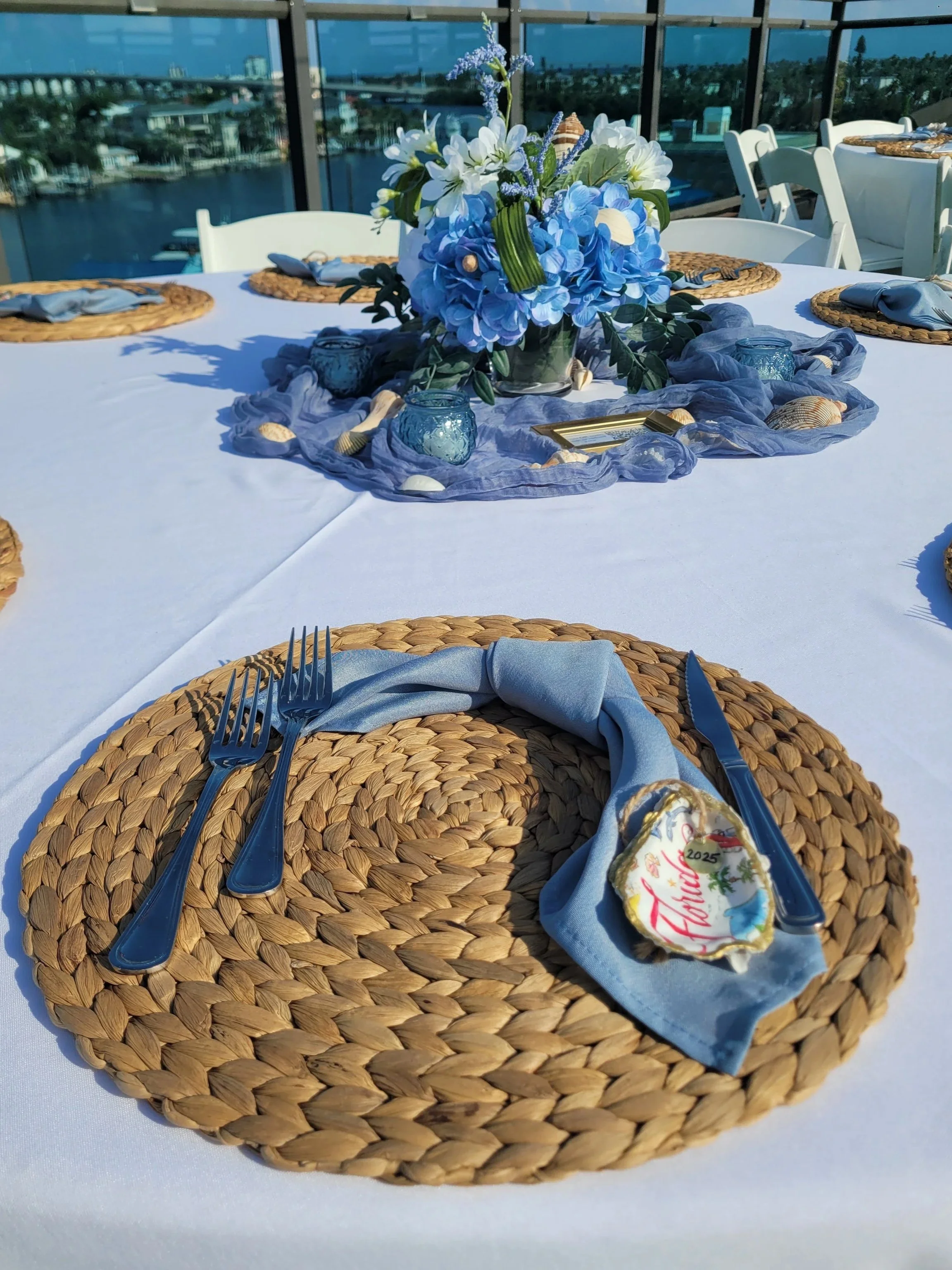 A table setting with a centerpiece of blue and white flowers, seashells, and small candles, arranged with a white tablecloth, woven placemats, and blue cloth napkins tied with golden rings, overlooking a waterfront view.