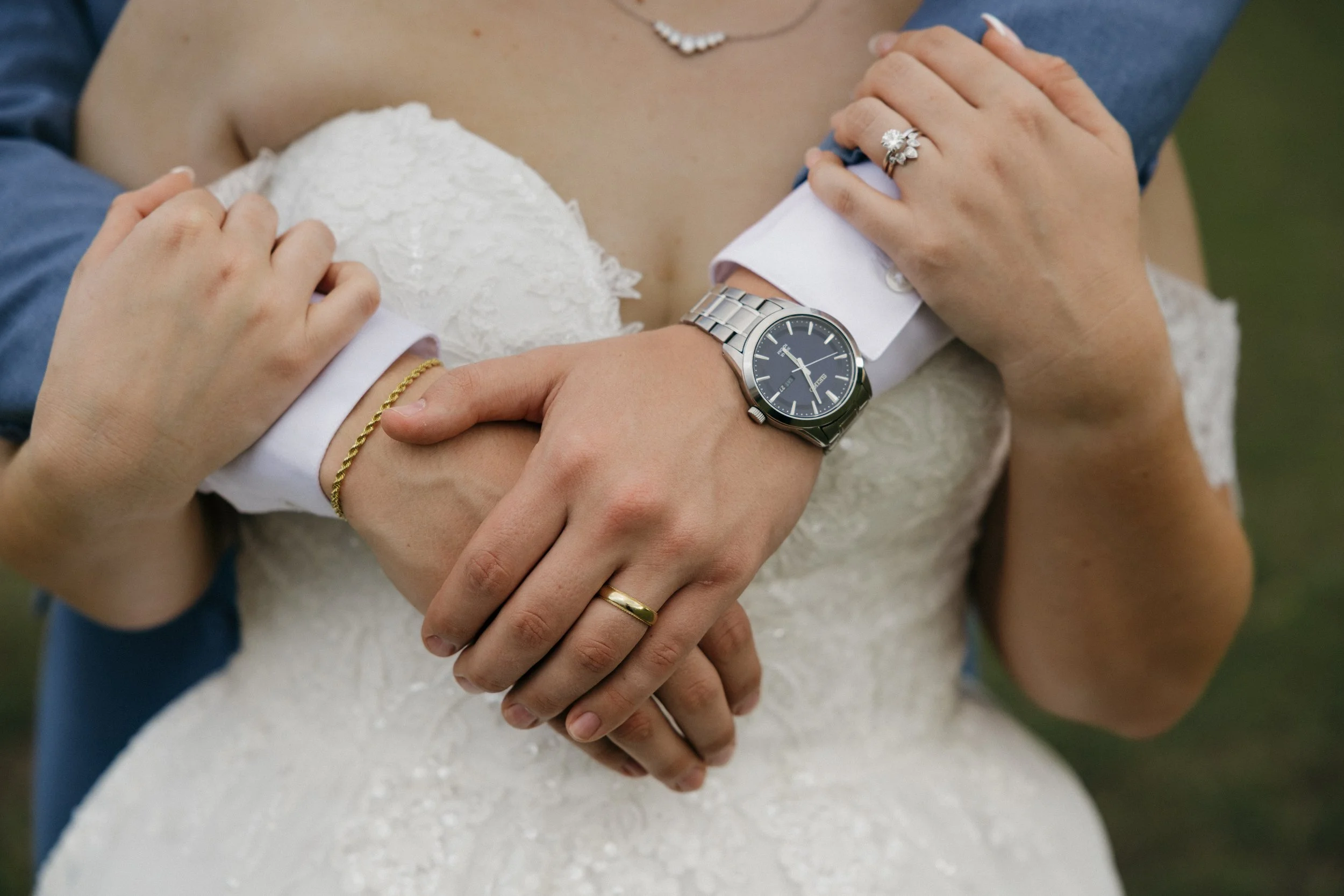 Close-up of a wedding couple's hands, with the bride's hand resting on the groom's chest. The groom wears a silver watch and a gold bracelet, while the bride has rings and a ring on her ring finger, and wears a lace wedding dress.
