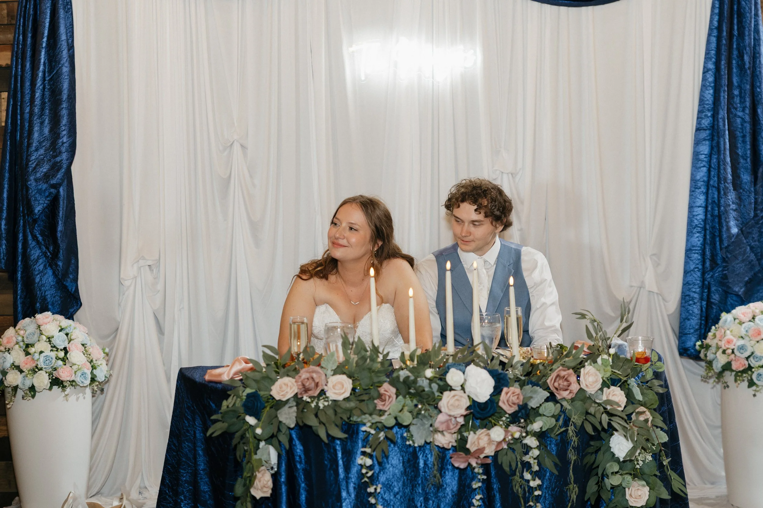 A bride and groom sitting at a decorated wedding reception table with floral arrangements and candles, in front of a white backdrop with blue curtains.