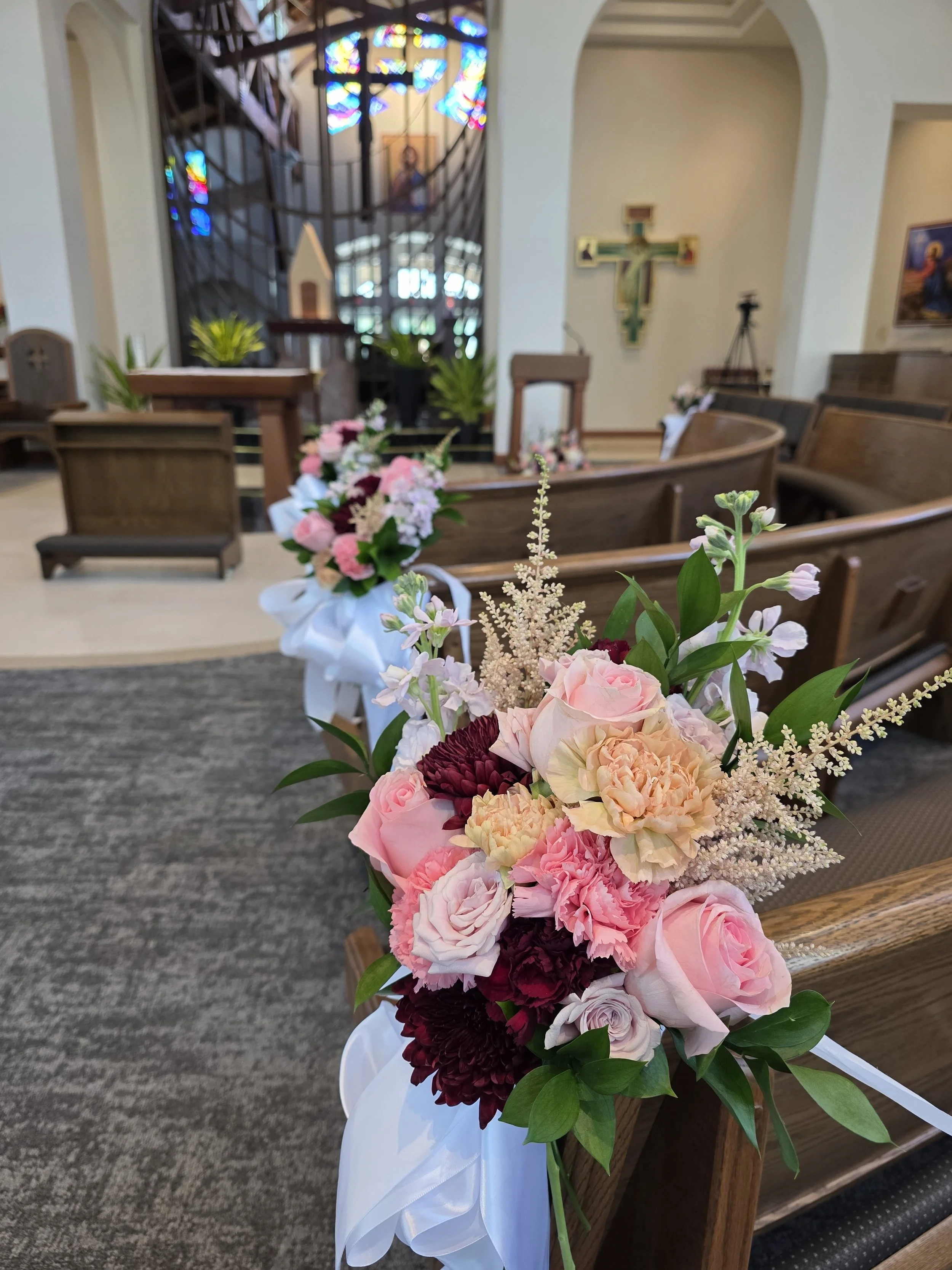 A floral arrangement with pink, white, and burgundy flowers and green leaves, decorated with a white ribbon, placed on a pew in a church or chapel interior with wooden pews, stained glass windows, and religious art in the background.
