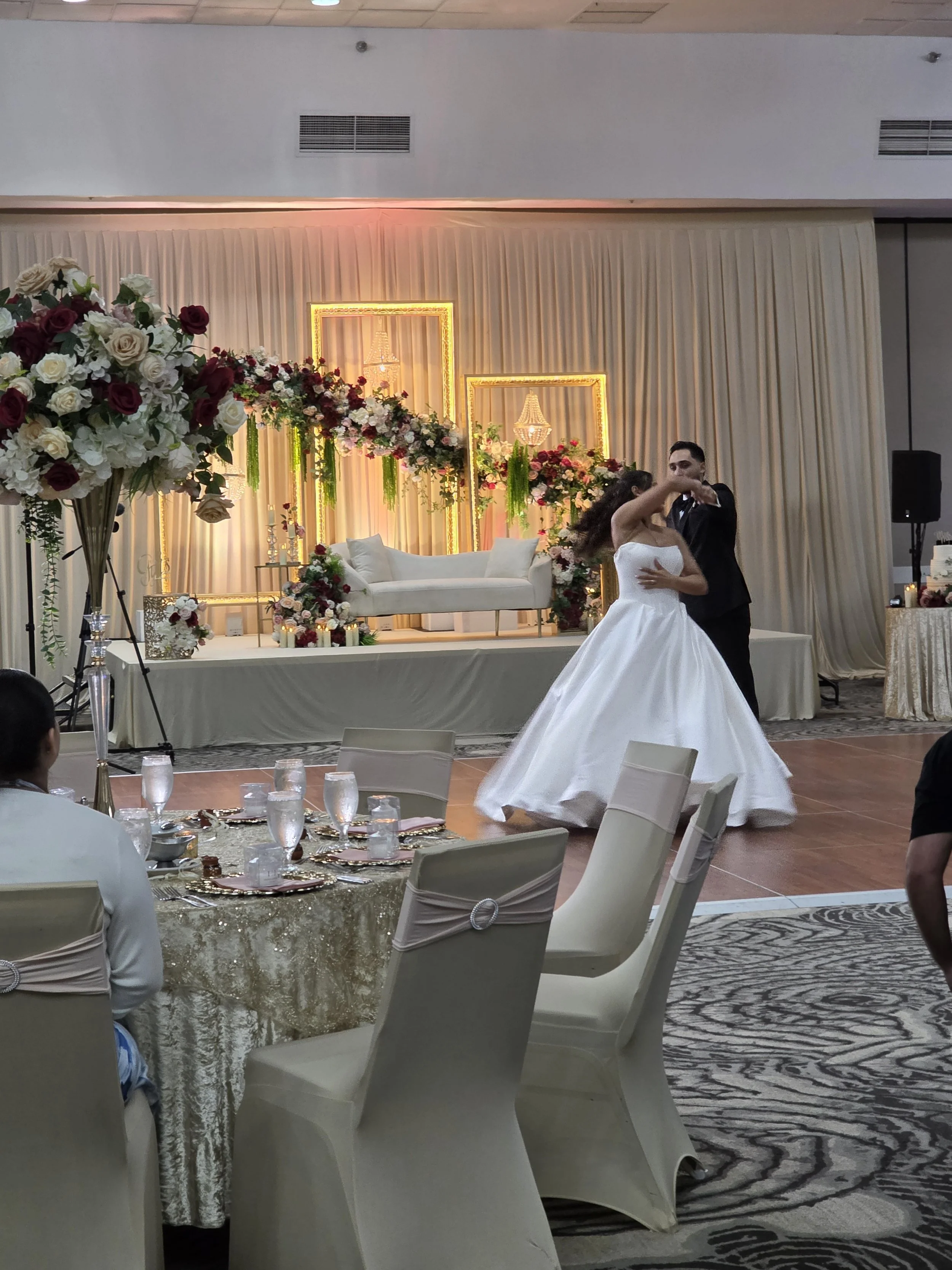 A bride and groom dancing at their wedding reception, with a decorated stage backdrop behind them featuring floral arrangements, chandeliers, and illuminated frames.