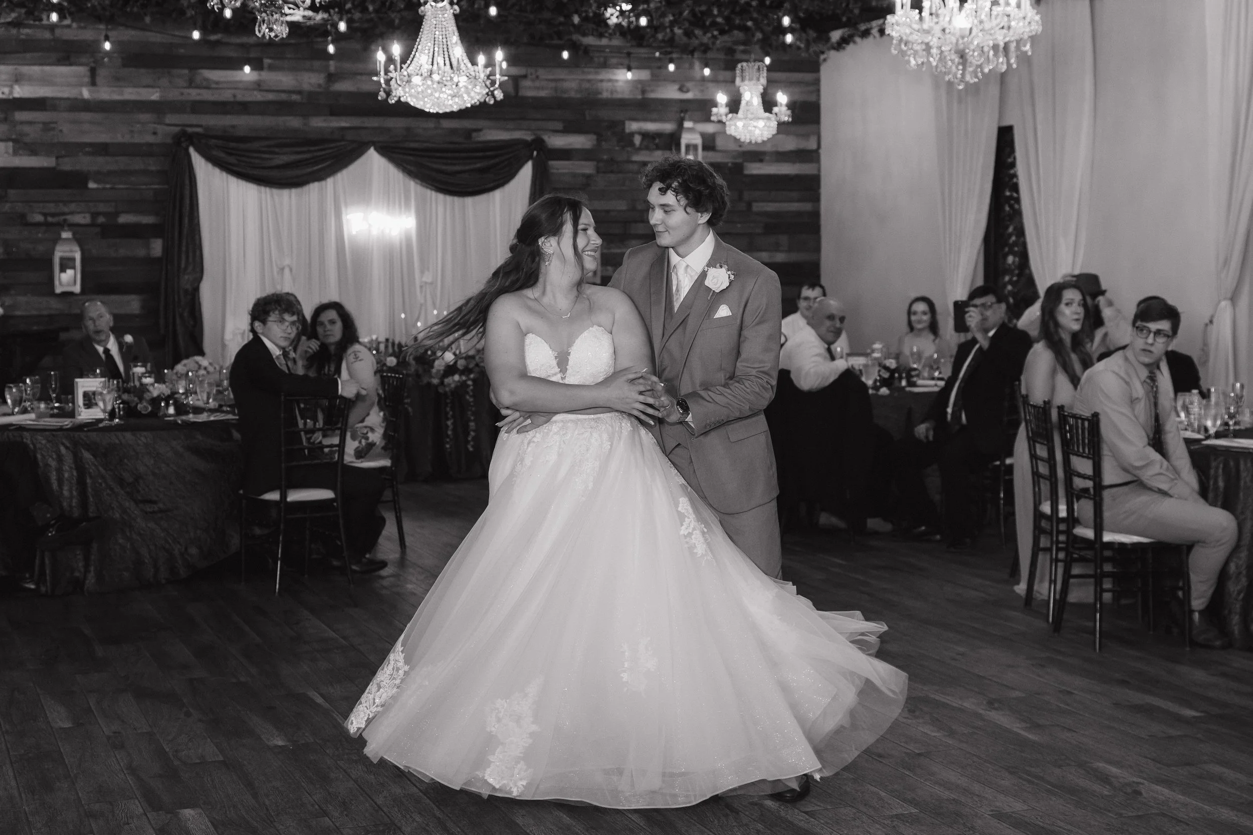 Wedding reception with bride and groom dancing, surrounded by seated guests, chandeliers, and rustic decor.