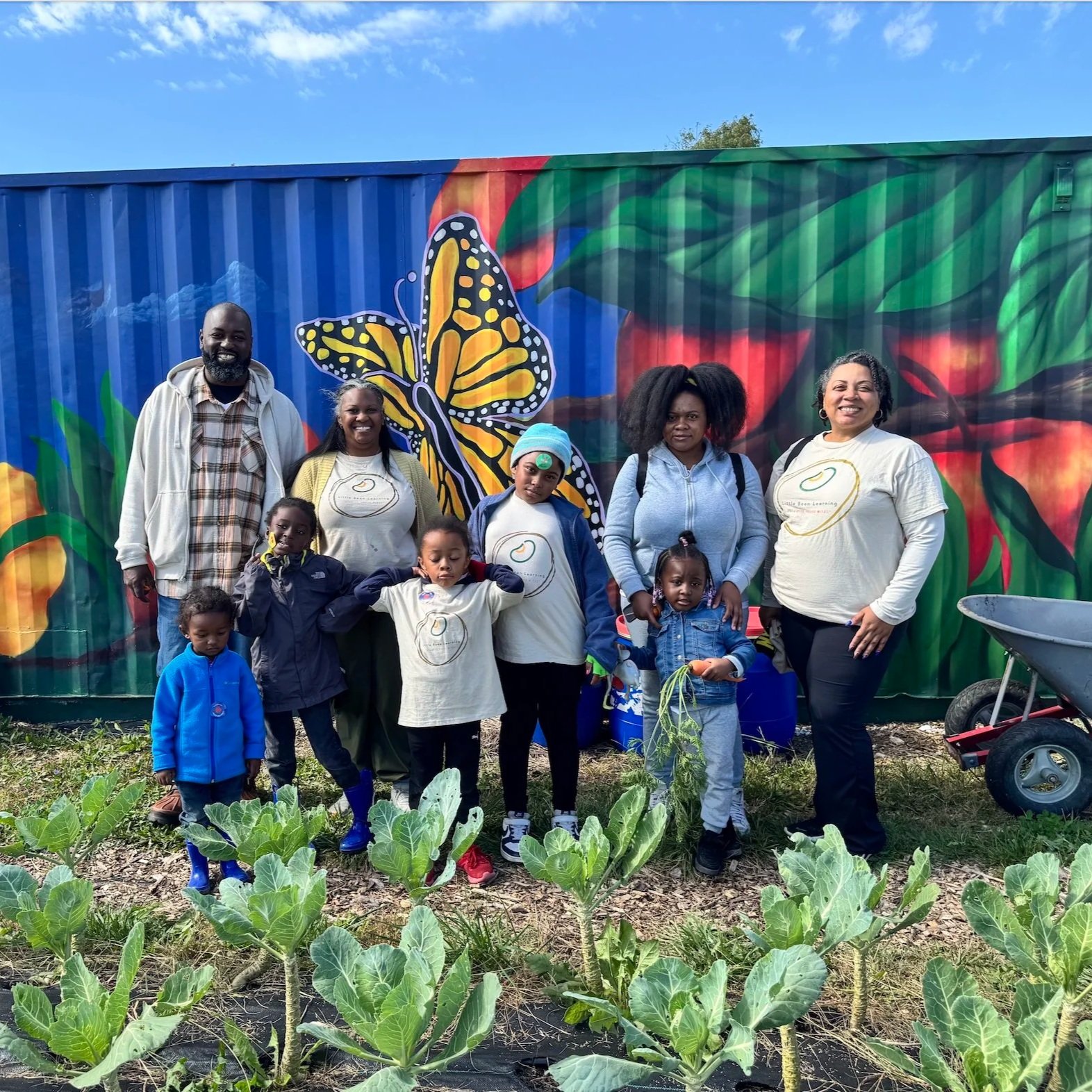Group of children and adults standing in front of a colorful mural of a butterfly and leaves, in a garden with leafy plants.