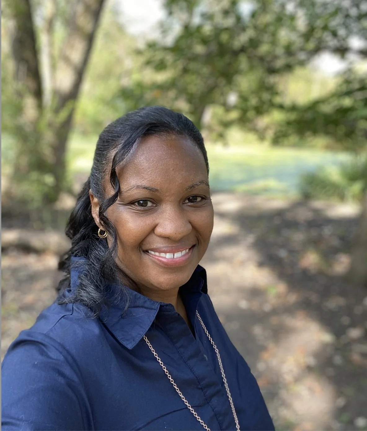 A woman smiling outdoors in a park or forested area, wearing a navy blue shirt and gold earrings, with trees and greenery in the background.