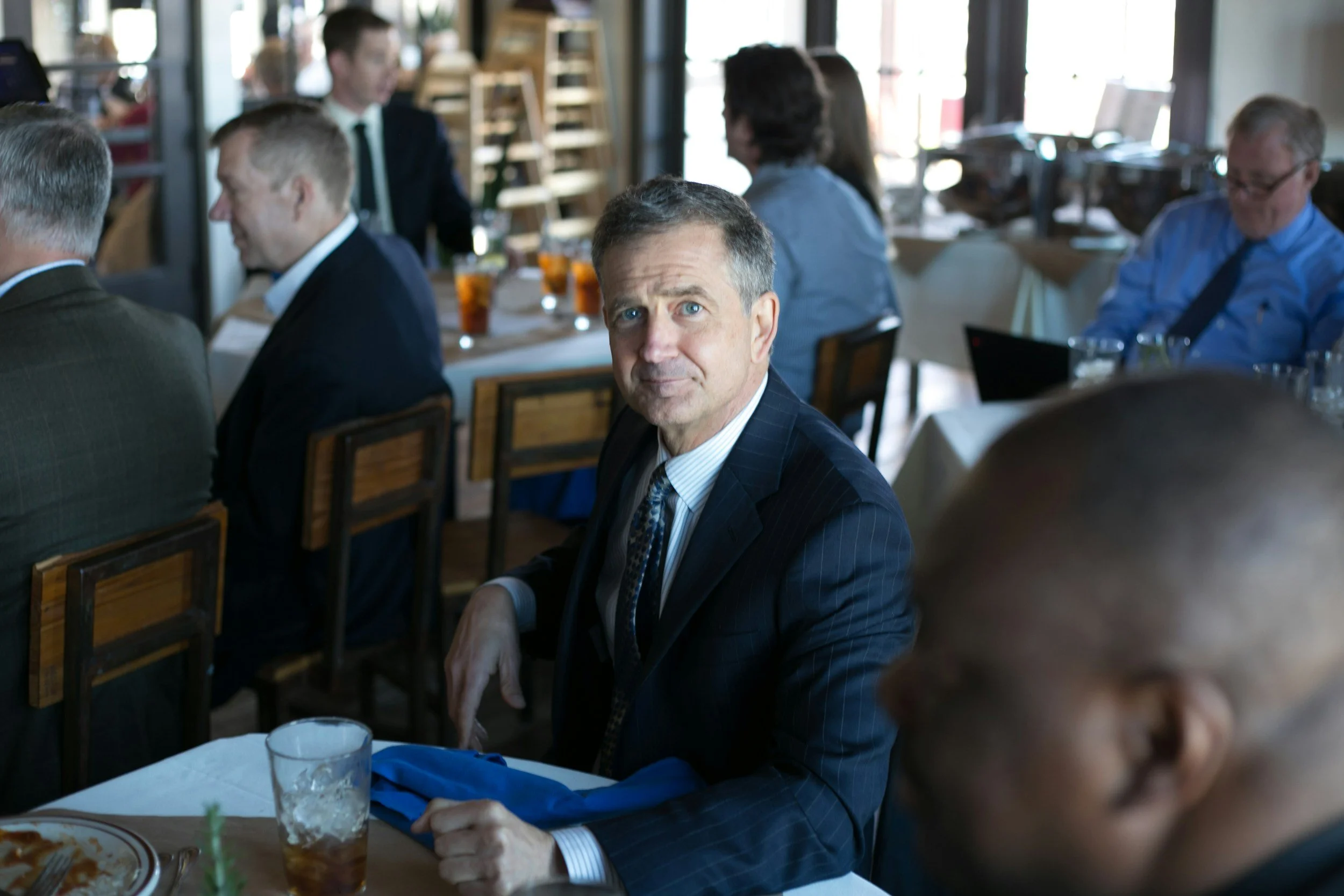 A man in a dark pinstripe suit and striped shirt sitting at a table in a restaurant, looking at the camera with blue eyes. Other middle-aged and older adults are seated at tables in the background, engaged in conversation, with drinks and food on the tables.