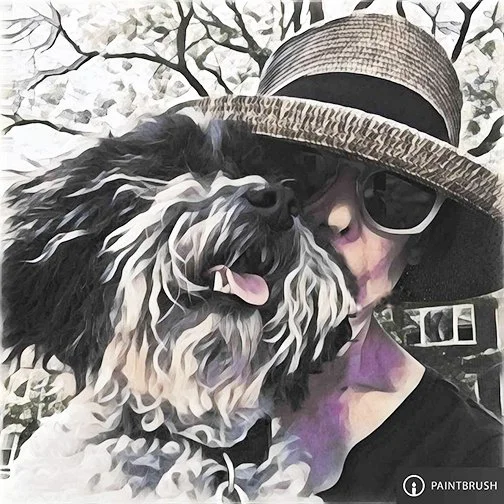 Person wearing a wide-brimmed hat and sunglasses, embracing a black and white dog outdoors with trees in the background.