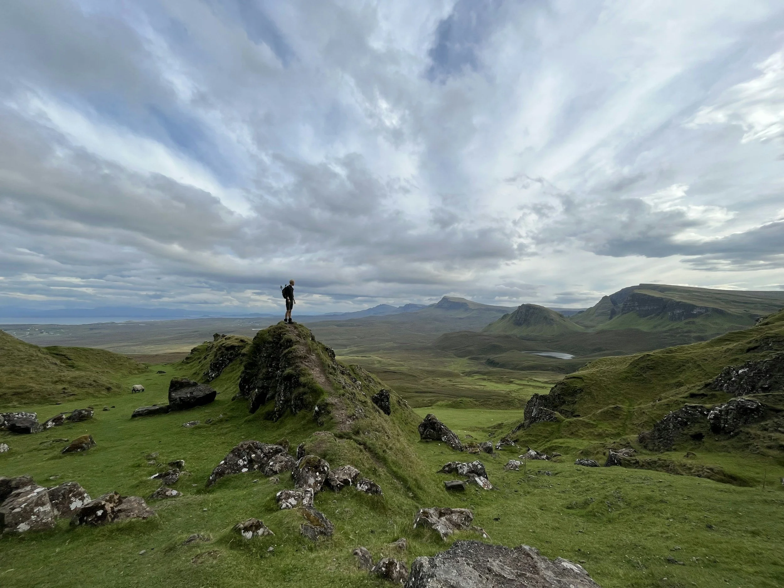 A lone hiker standing on a rocky hilltop in a vast green landscape under a cloudy sky, with mountains and a lake in the distance.