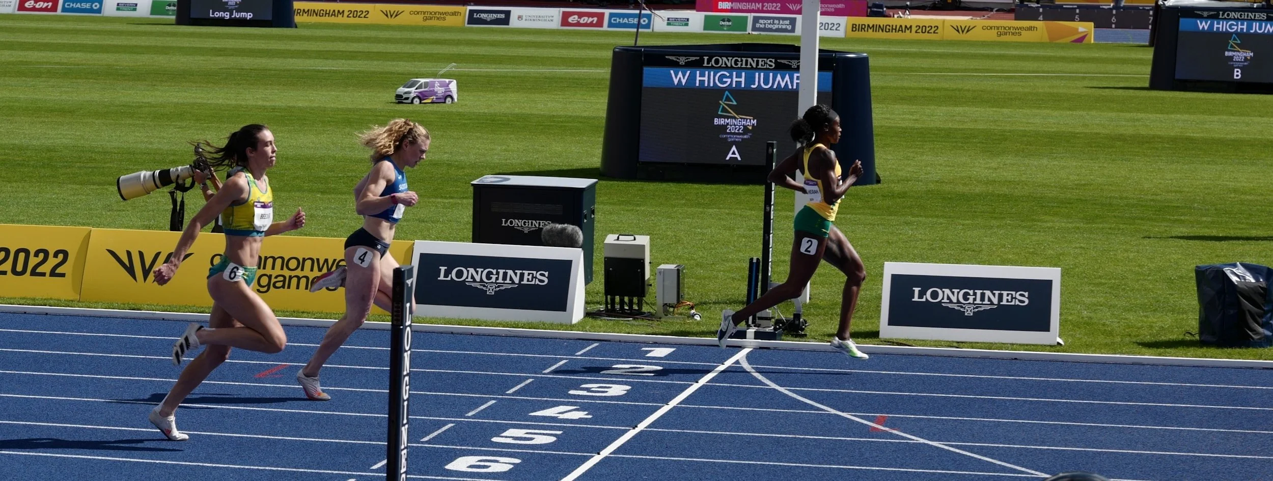 Three female athletes finish a race on a blue track, with one runner in the lead near the finish line, and the other two close behind, during the Birmingham 2022 Commonwealth Games.